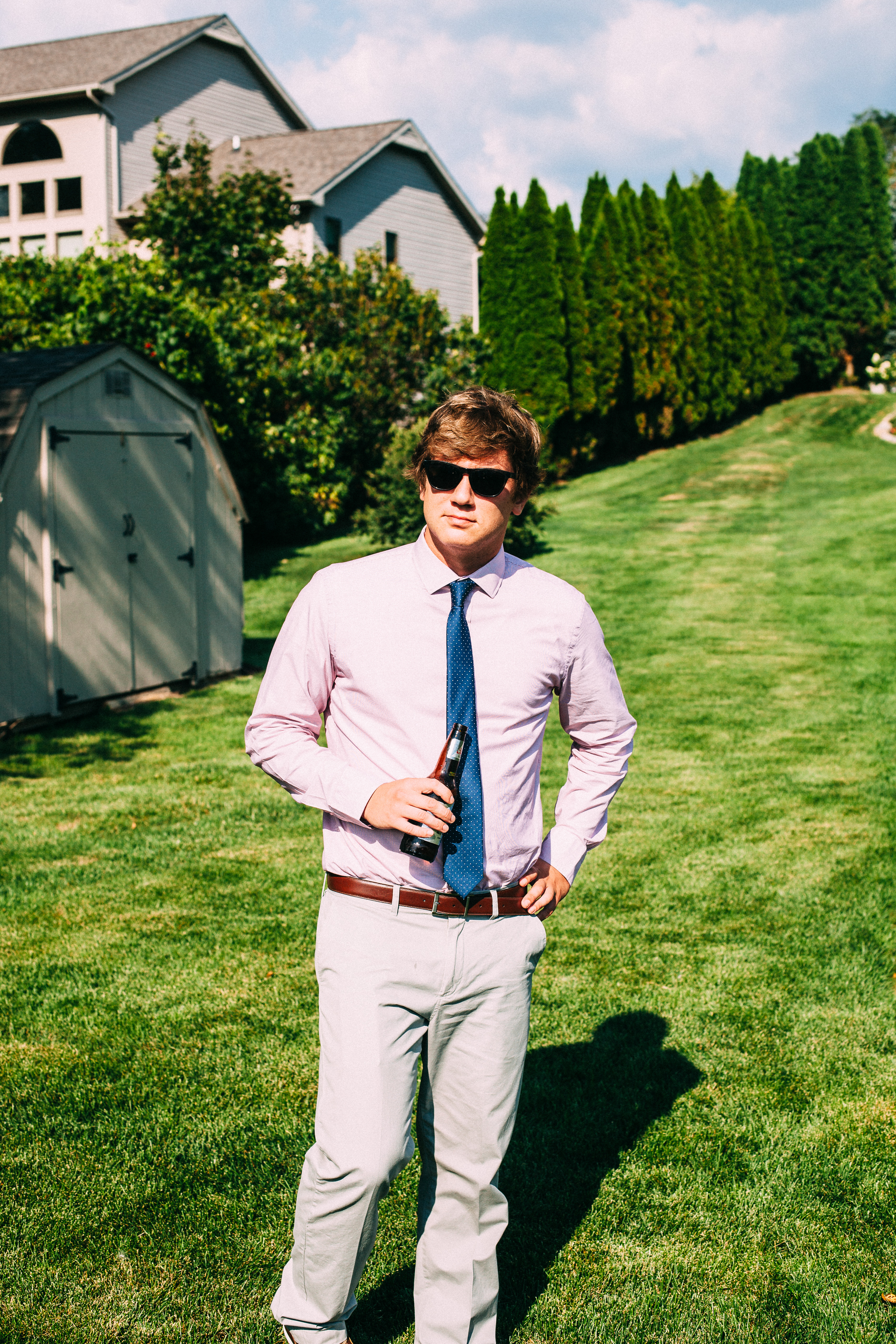 Groomsman in sunglasses, pink shirt, and blue dotted tie standing on a sloping lawn by a garden shed.