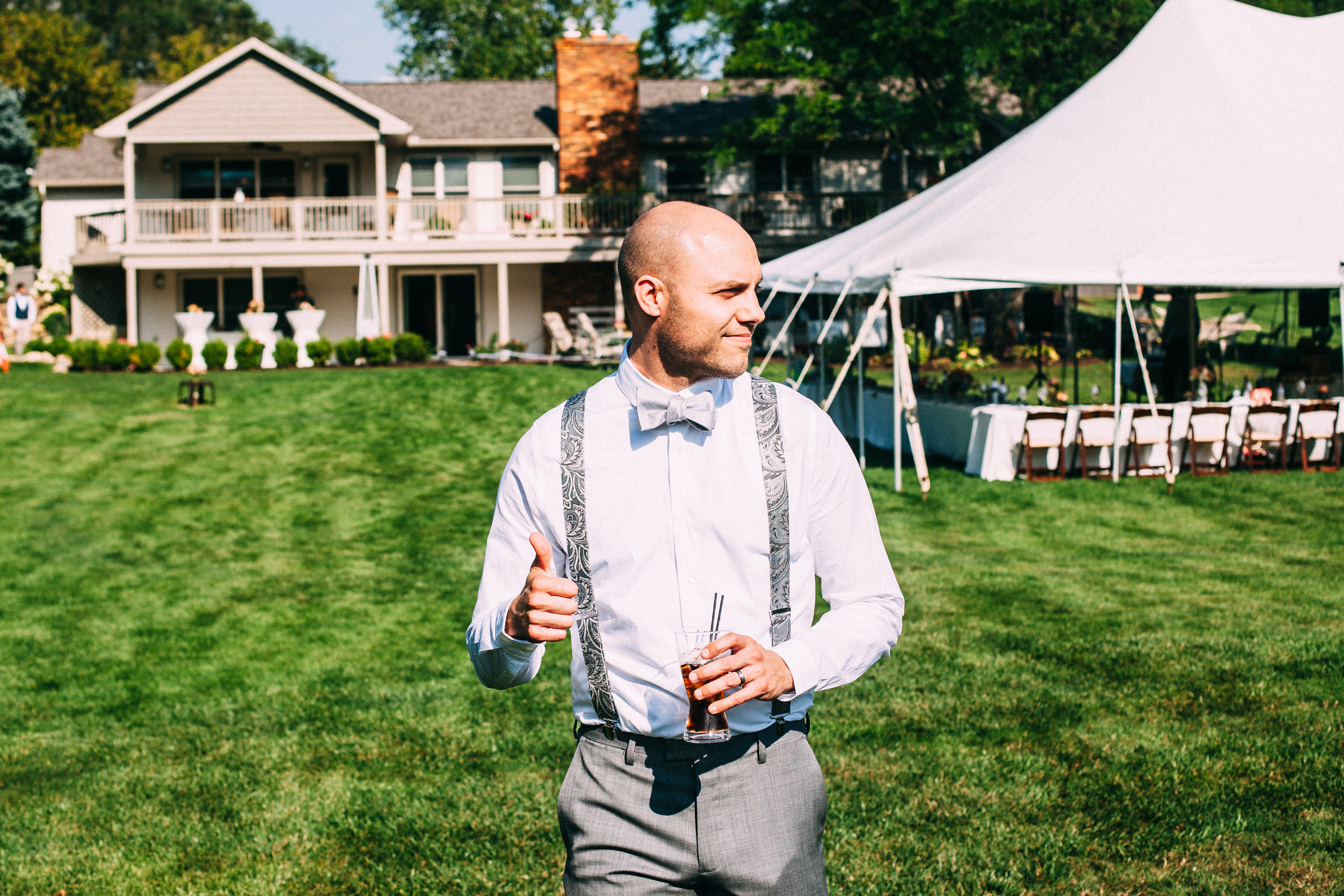 Groomsman in patterned suspenders and bow tie giving a thumbs up with a cocktail on a sunny lawn.