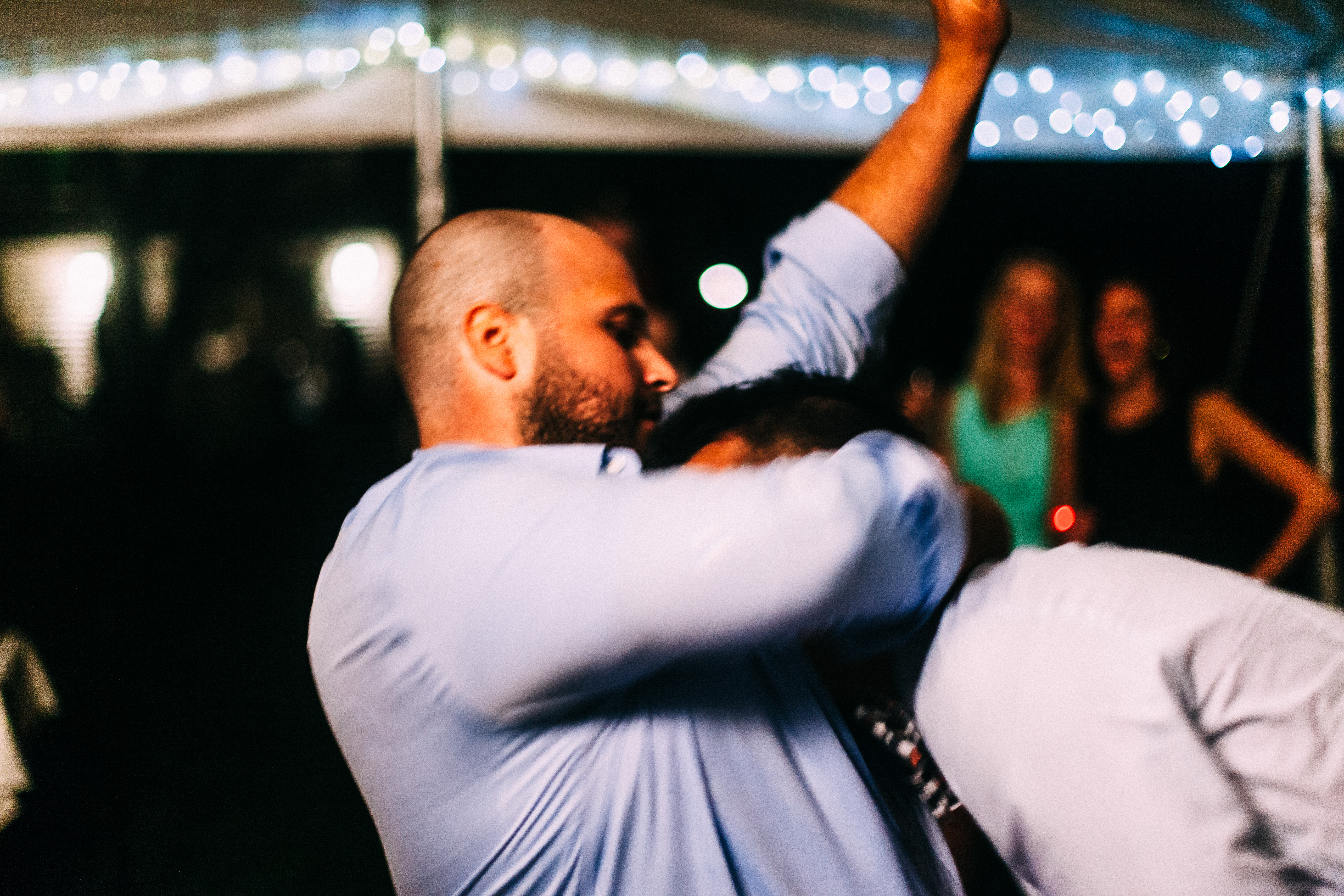 Two men dancing excitedly and bumping shoulders under reception tent string lights with guests behind.