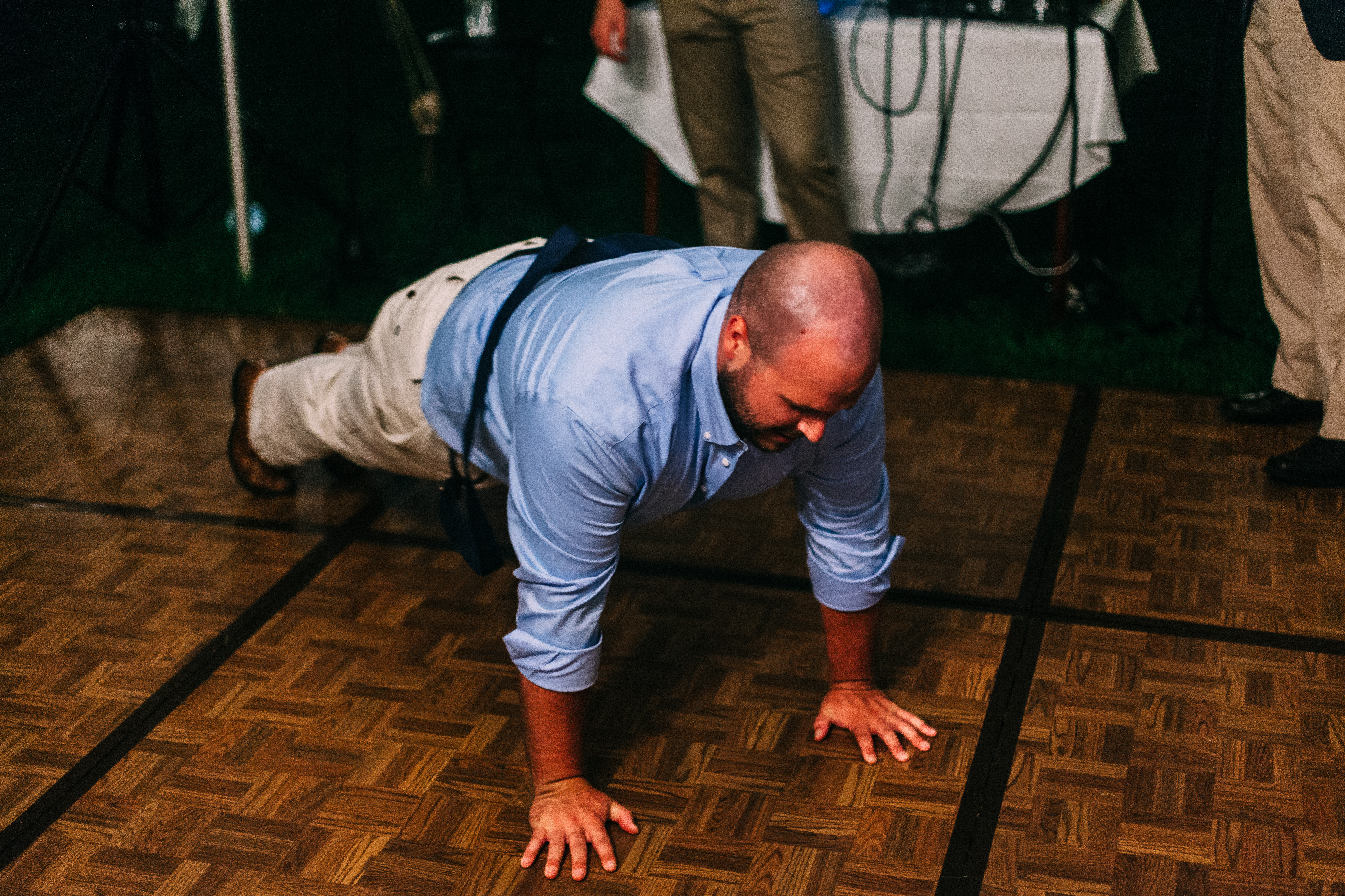 Man in blue shirt doing pushups on the wooden parquet dance floor at a nighttime wedding reception.