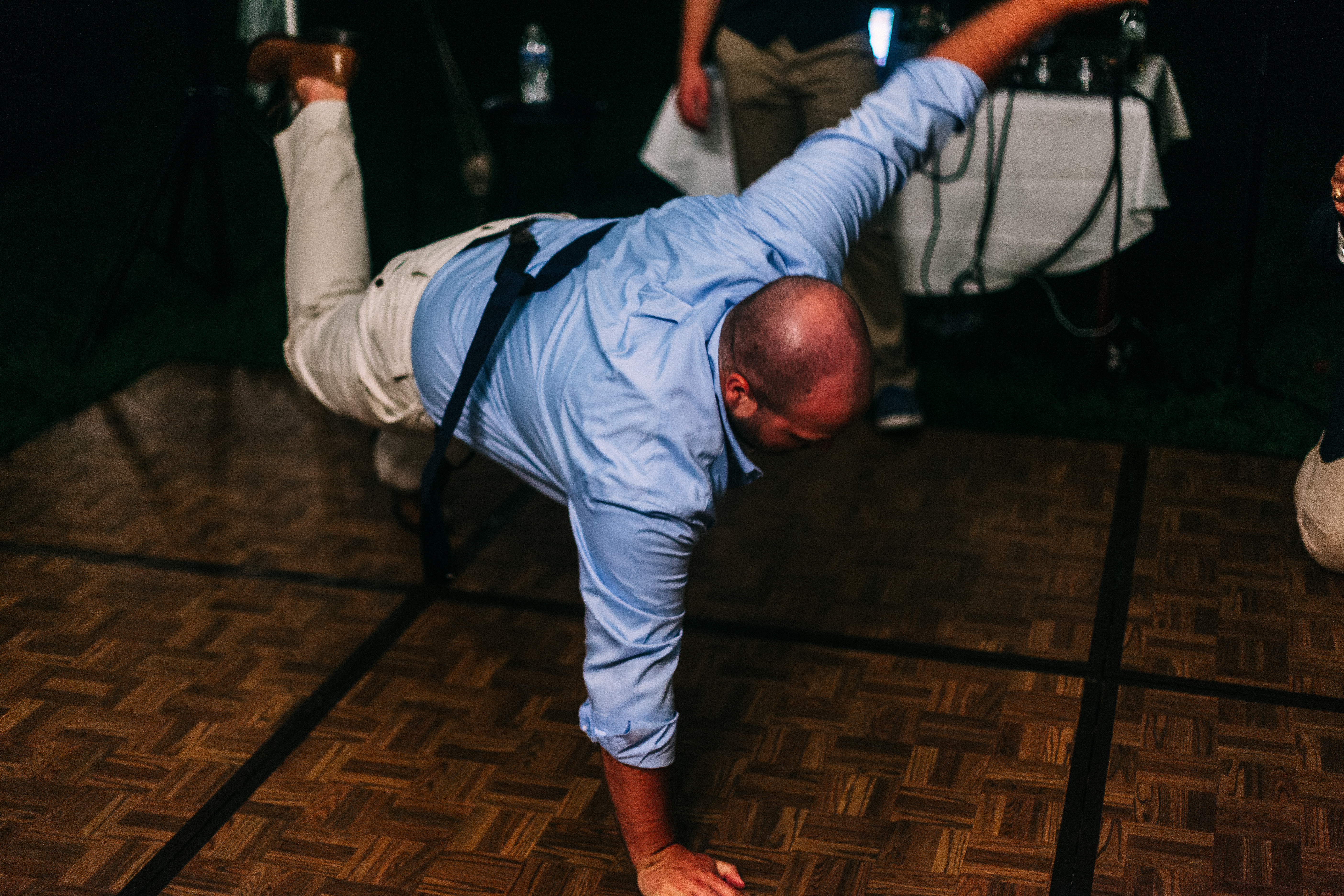 Man in suspenders performing a one-handed dance freeze on the wooden parquet floor at a wedding party.