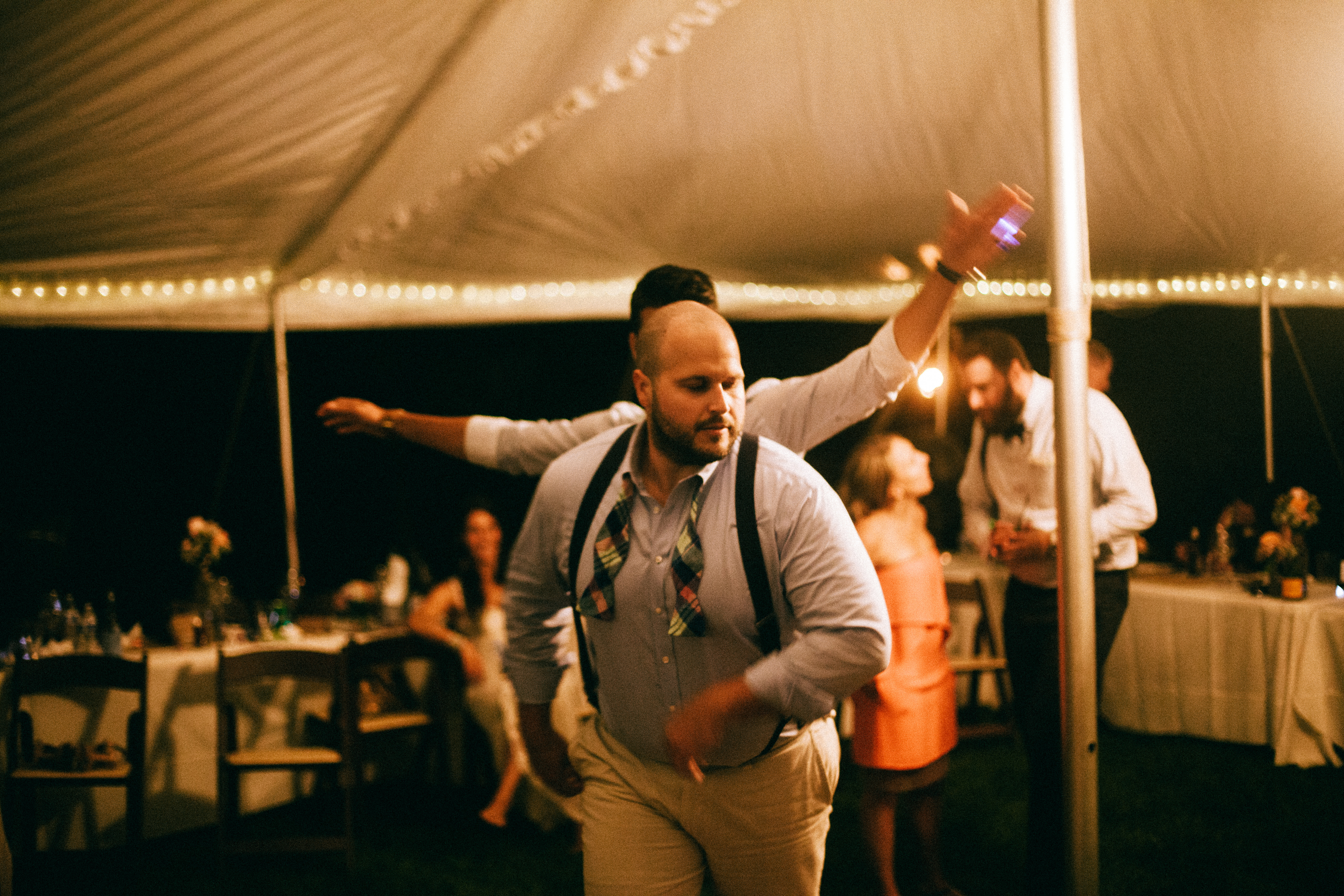 Bald man in suspenders dancing animatedly across the floor under tent lighting with arms outstretched.