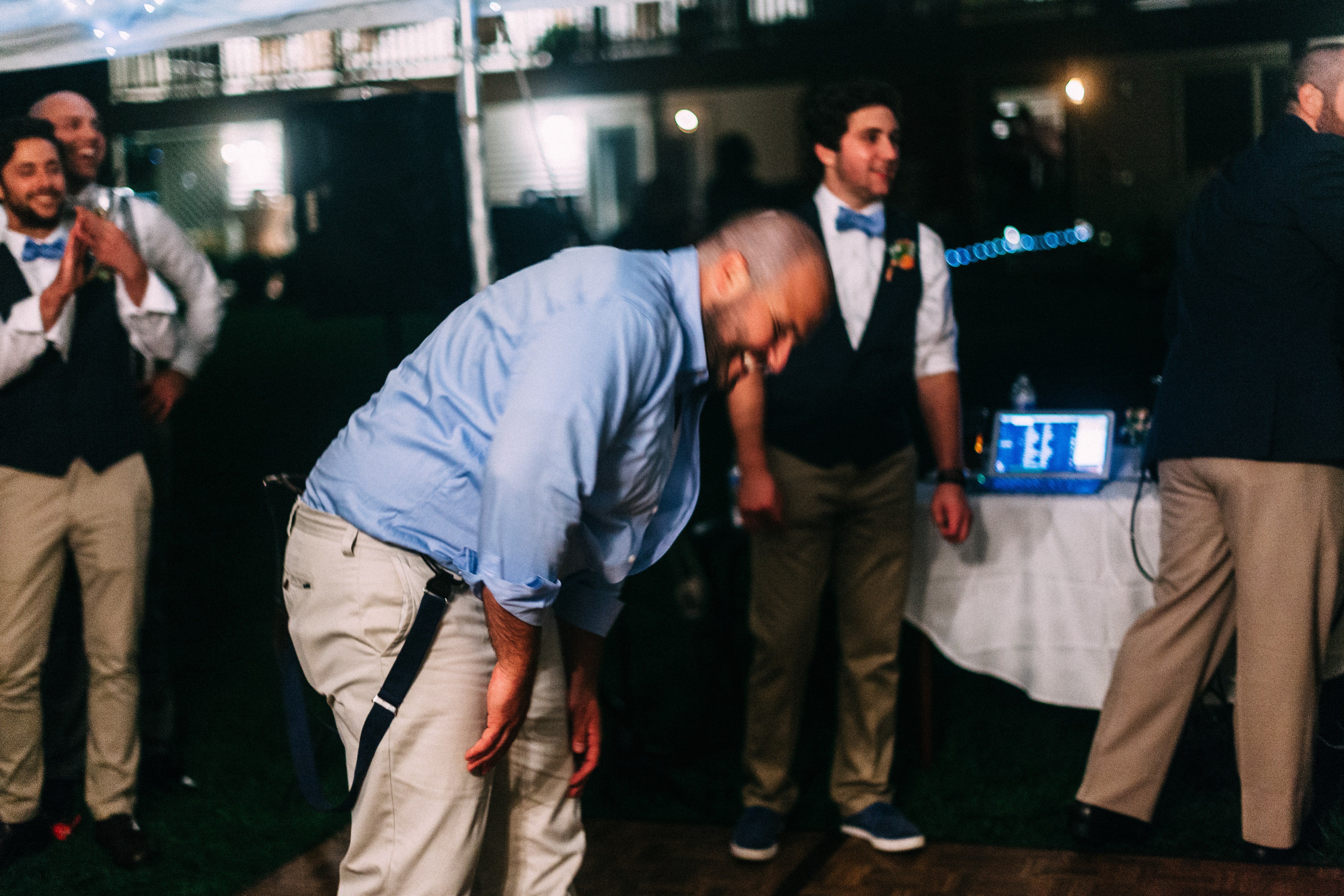 Man in blue shirt laughing while bowing down on the dance floor surrounded by smiling groomsmen.