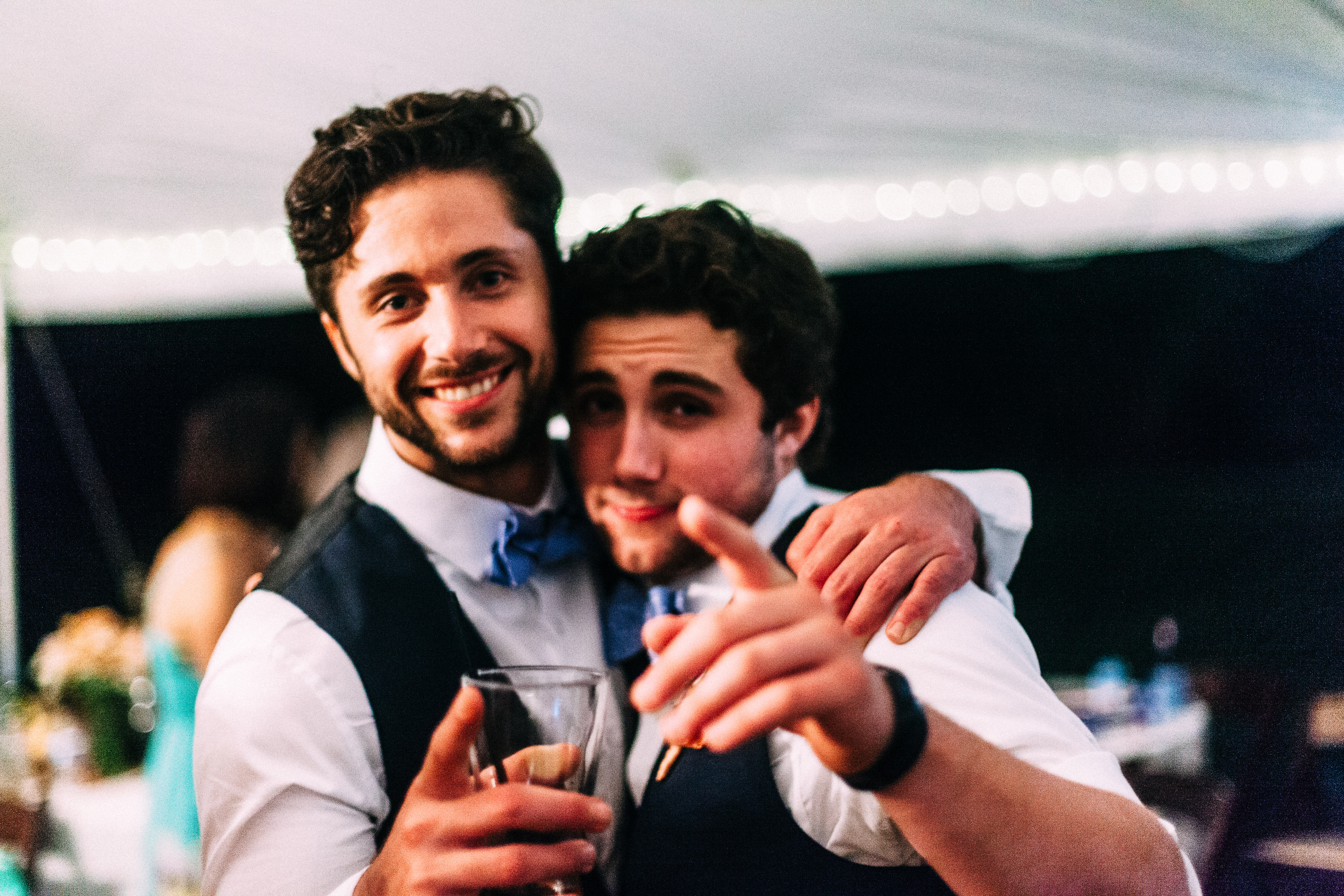 Two smiling groomsmen in matching navy vests and blue bow ties with arms around each other at a reception party, with one pointing playfully toward the camera while holding a cocktail, shot with a Canon 5D Mark II.