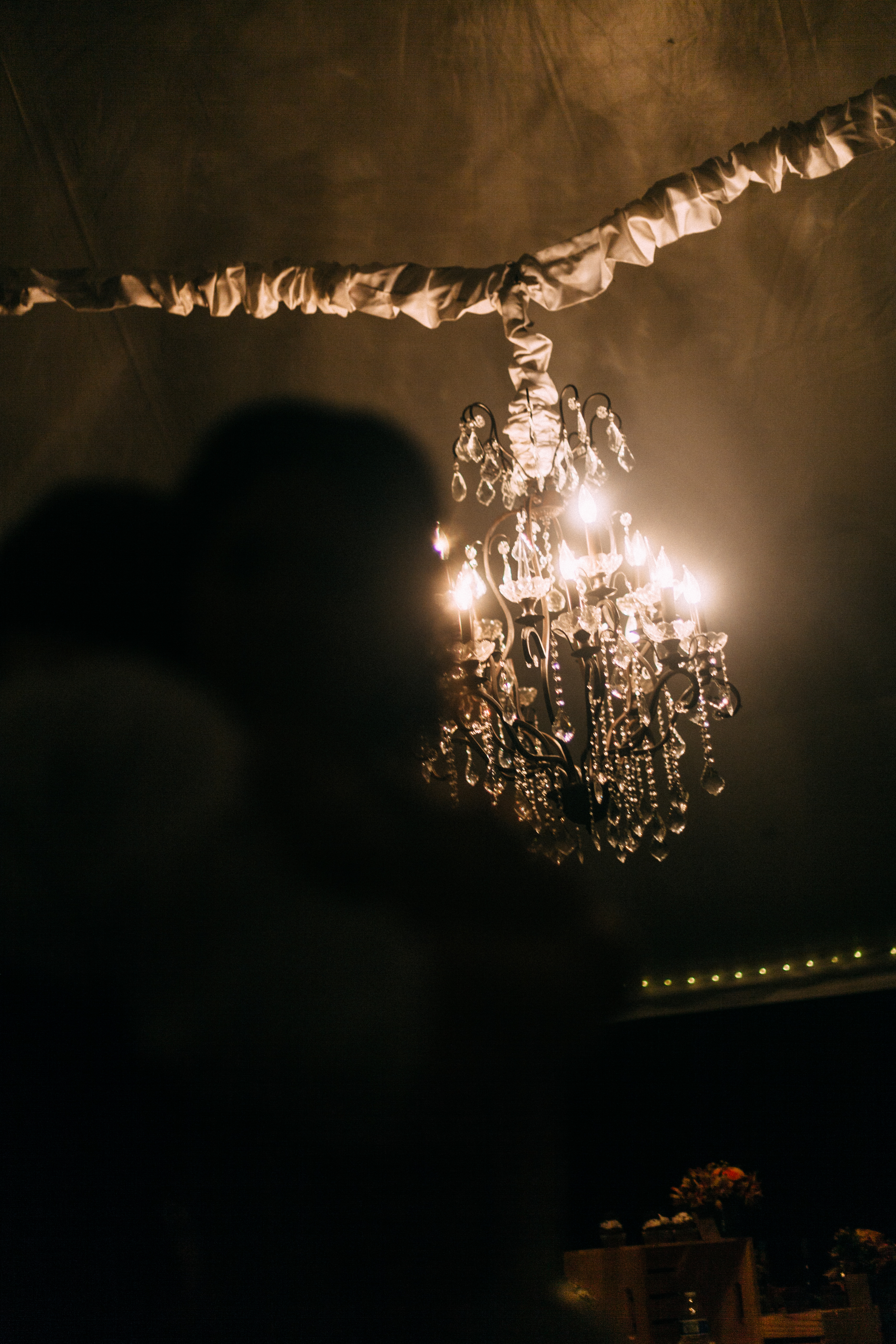 An illuminated crystal chandelier hanging beneath gathered fabric draping inside a dark wedding reception tent at night, shot with a Canon 5D Mark II.