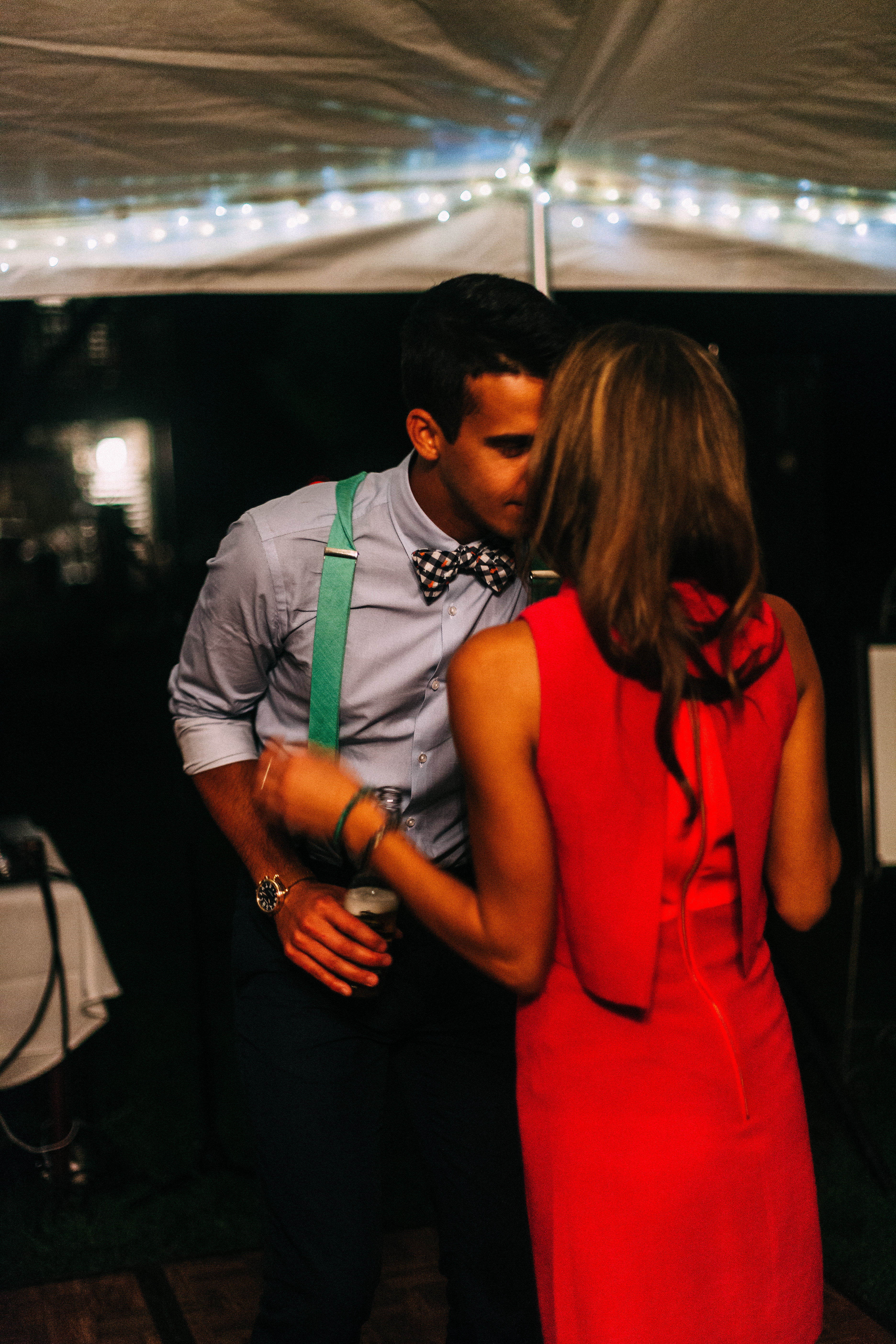 A groomsman in light blue shirt, green suspenders, and bow tie whispering in the ear of a woman in a sleeveless red dress on the dance floor at night, shot with a Canon 5D Mark II.