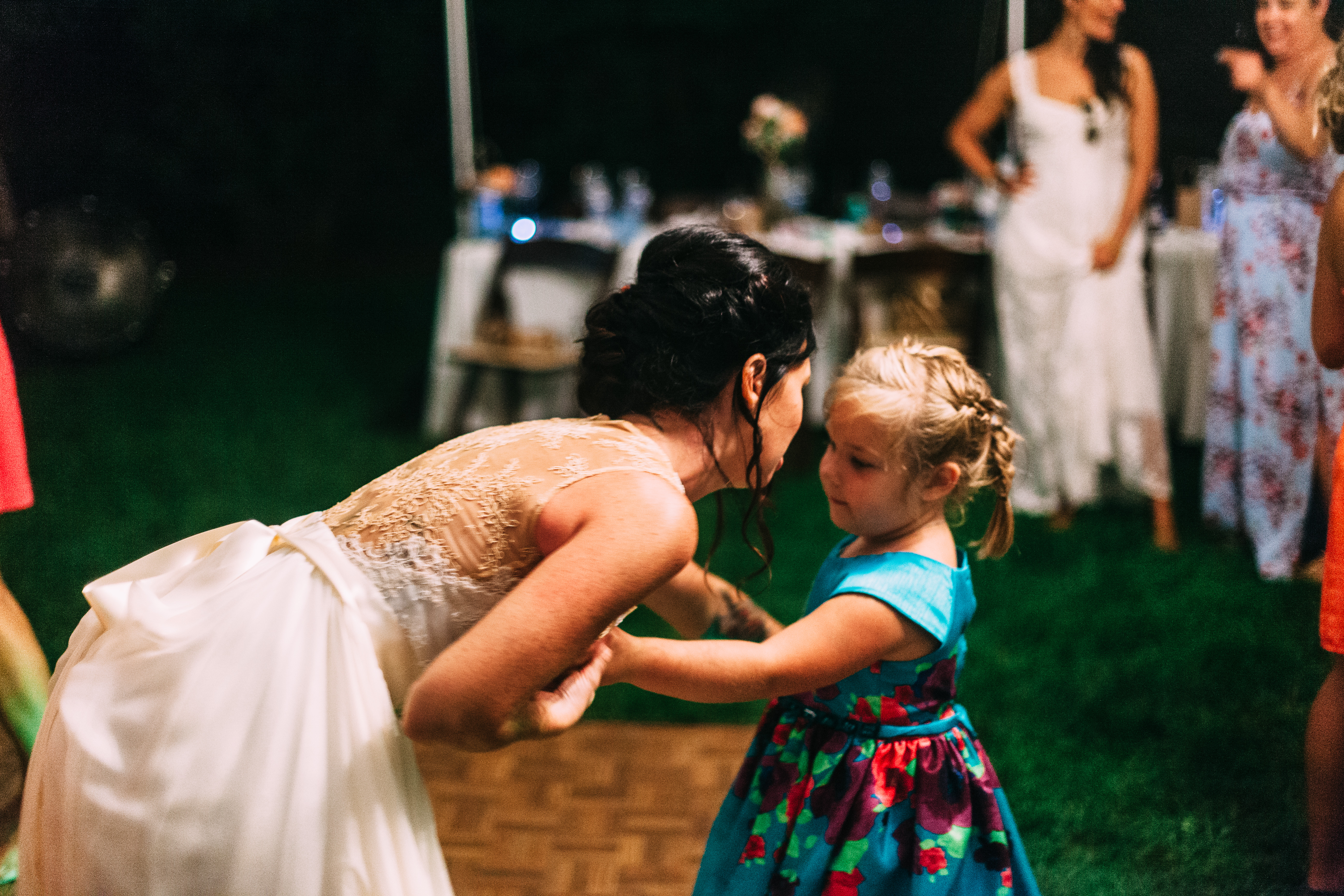 The bride in a lace gown leaning down smiling to dance hand-in-hand with a little girl in a vibrant floral dress on the reception dance floor, shot with a Canon 5D Mark II.