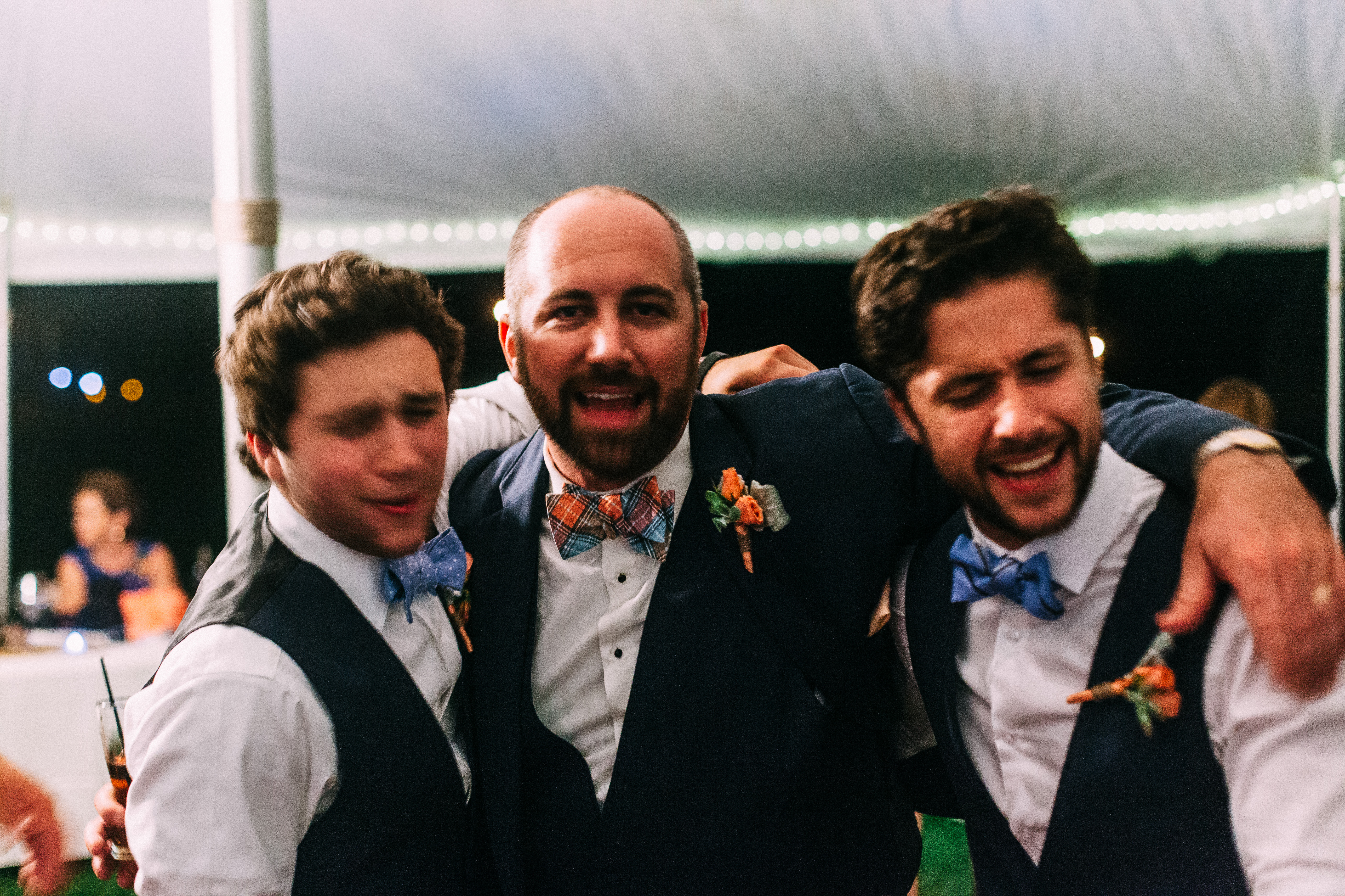 The groom in a navy blazer and bow tie singing passionately with arms around two smiling groomsmen on the reception dance floor at night, shot with a Canon 5D Mark II.