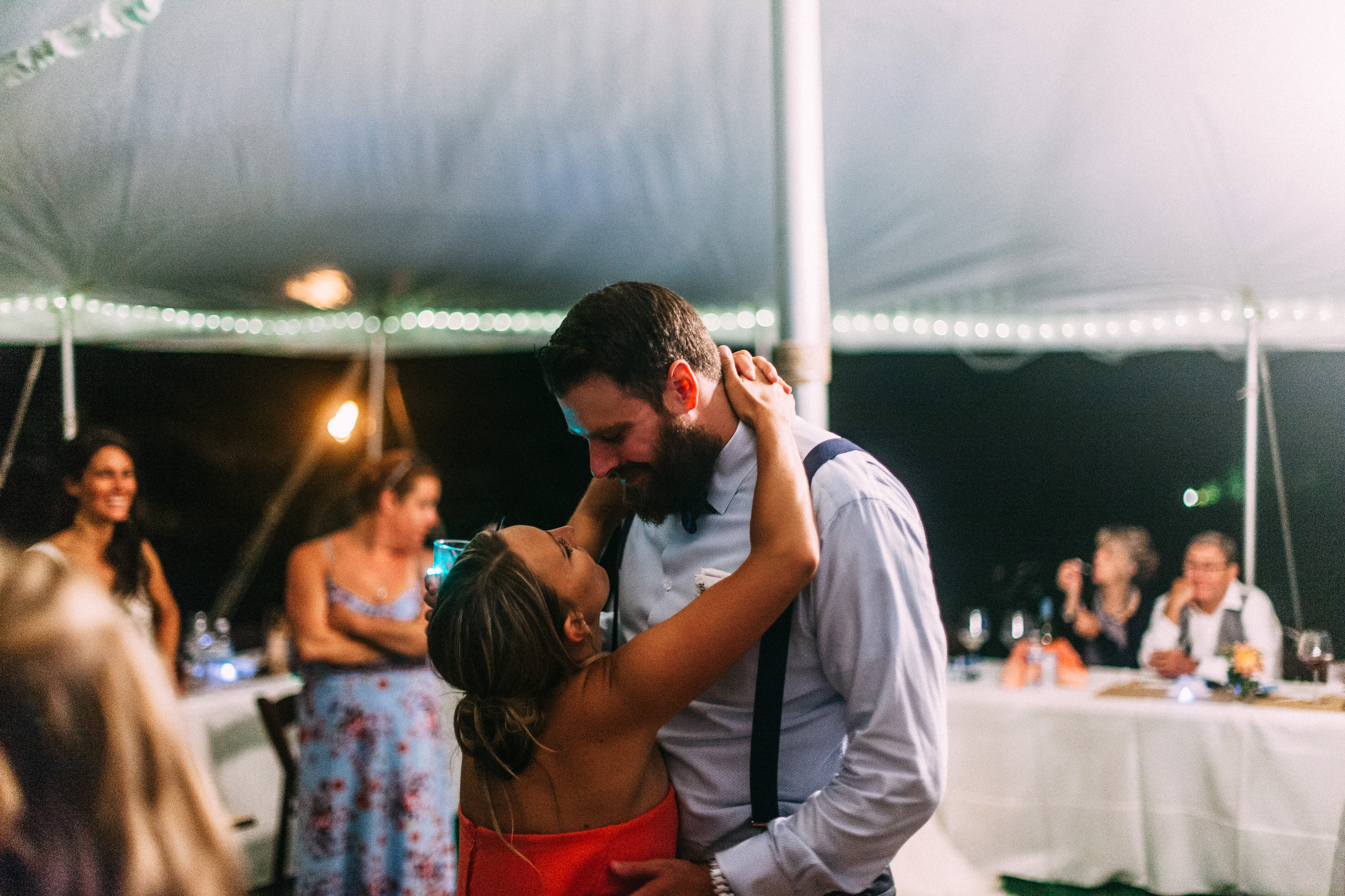A woman in a coral red dress with arms wrapped around the neck of a bearded man in a light blue dress shirt and navy suspenders as they slow dance together under a tent with string lights at a nighttime wedding reception, shot with a Canon 5D Mark II.