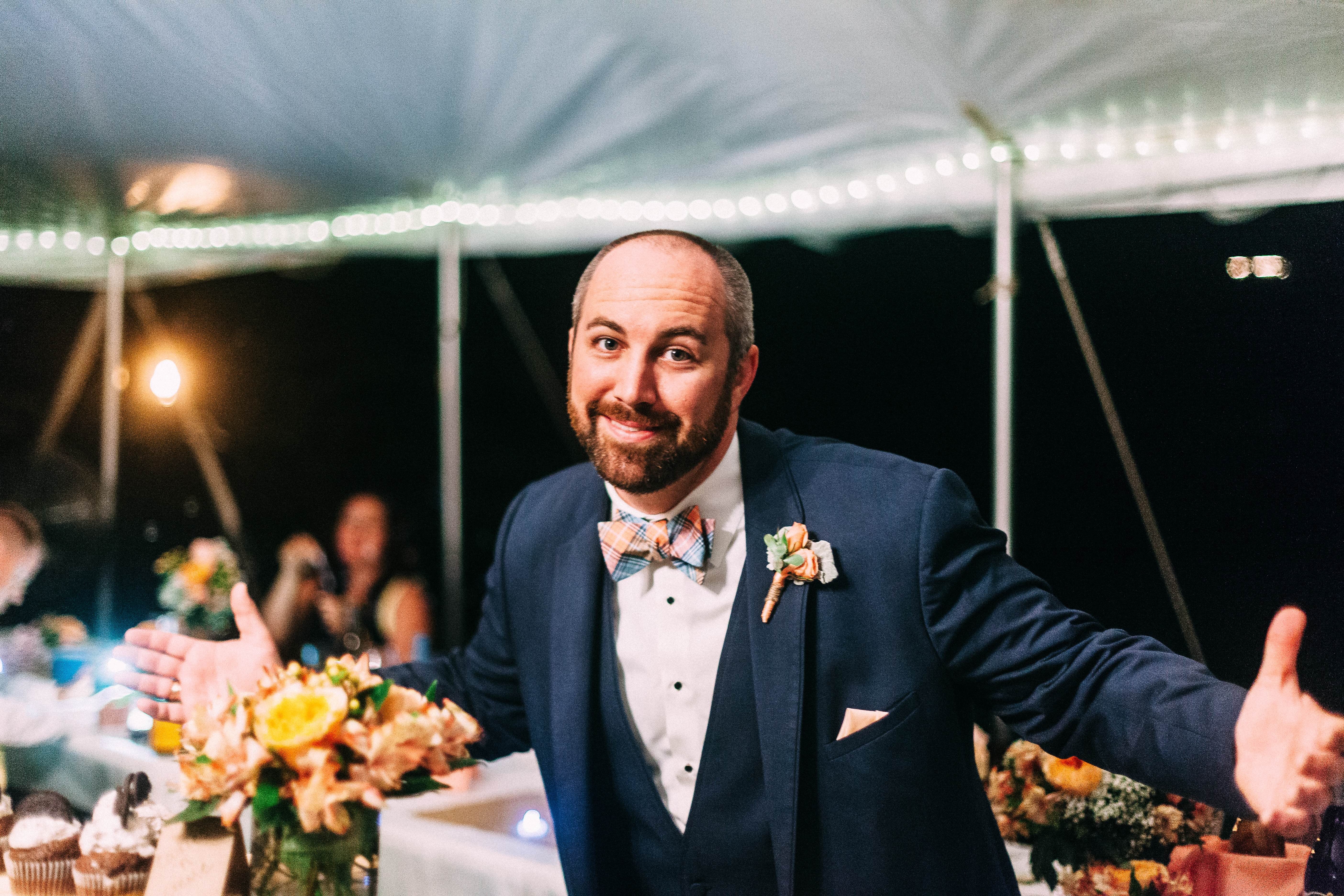 The smiling groom in a navy suit, plaid bow tie, and floral boutonniere shrugging with arms outstretched wide above reception dinner tables at night, shot with a Canon 5D Mark II.