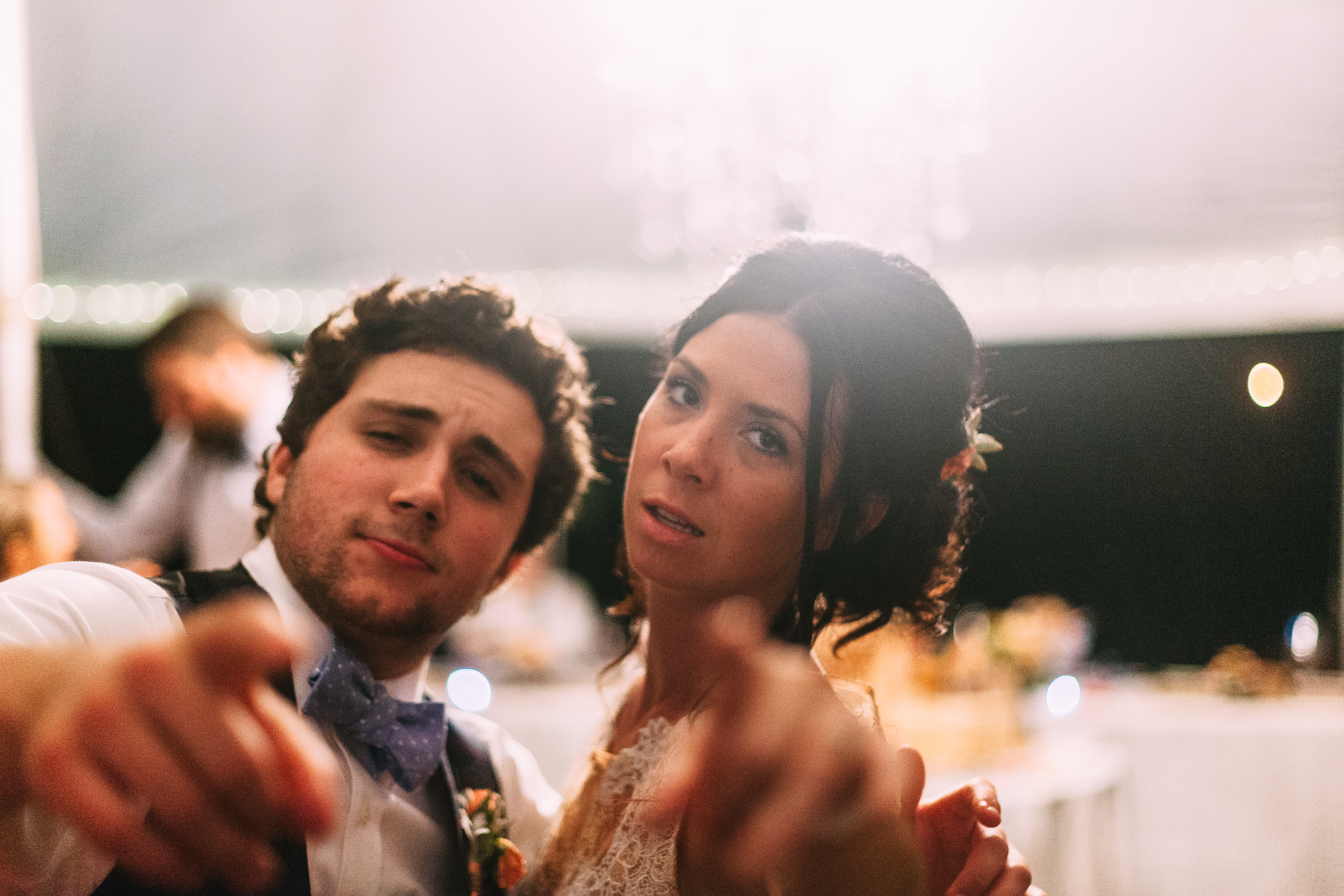 The bride in a lace wedding dress and a man with curly hair and blue bow tie pointing directly forward toward the camera with playful expressions under bright reception lighting at night, shot with a Canon 5D Mark II.