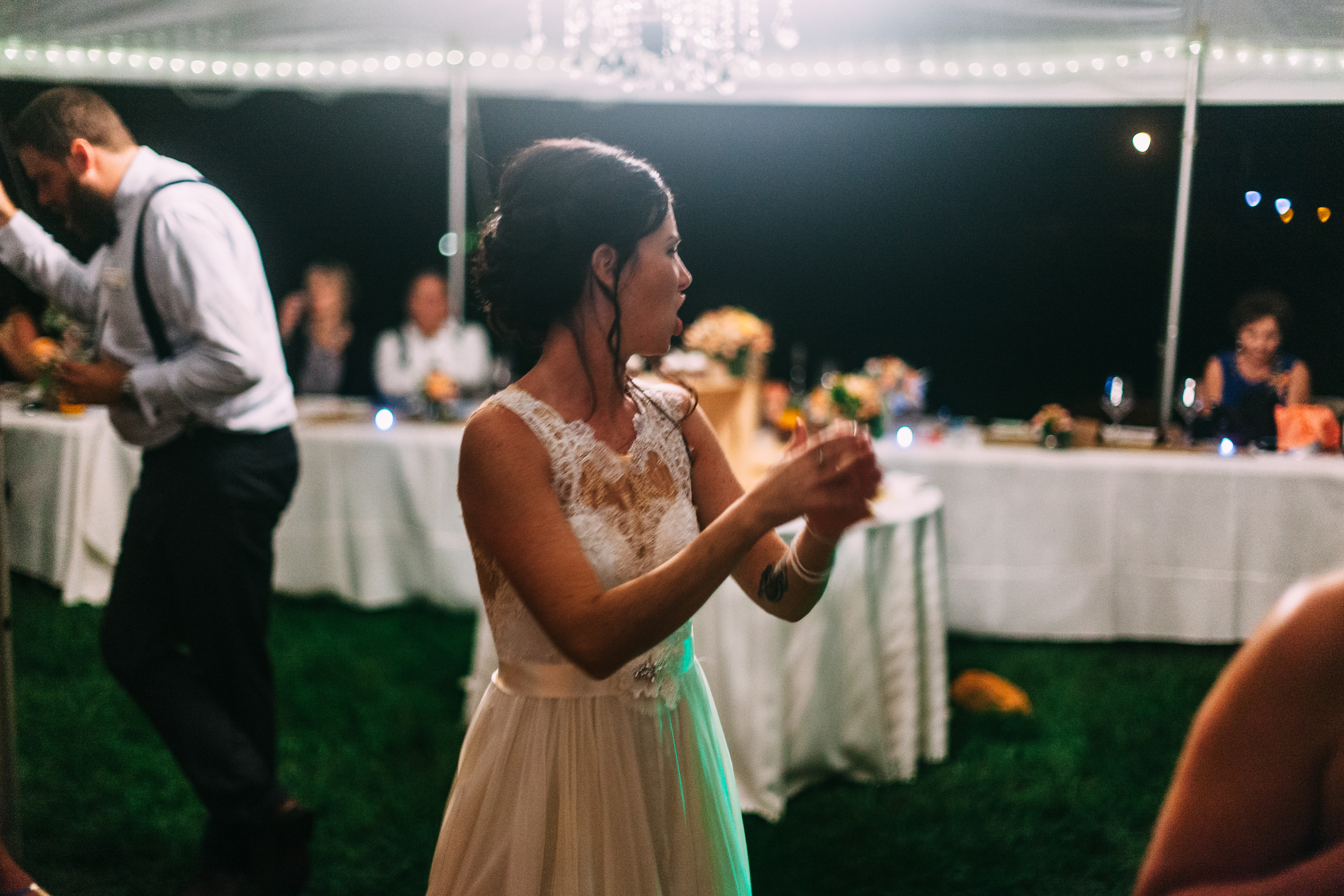 The bride in a sleeveless lace wedding dress clapping her hands enthusiastically on the lawn dance floor while guests dance around her under a reception tent at night, shot with a Canon 5D Mark II.