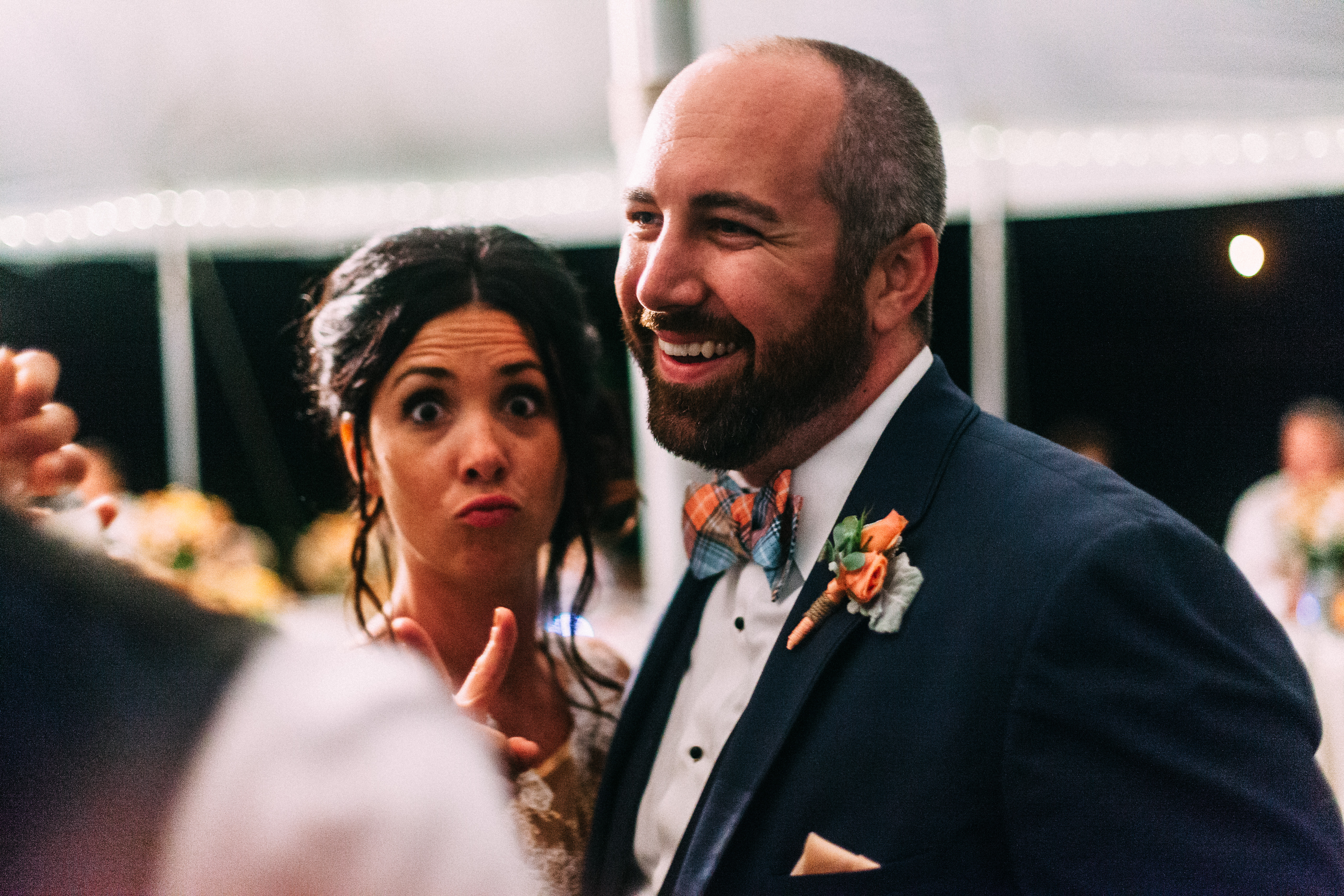 Close portrait of the smiling bearded groom in his navy suit and plaid bow tie while the bride playfully makes a funny wide-eyed face and peace sign behind his shoulder at their wedding reception, shot with a Canon 5D Mark II.