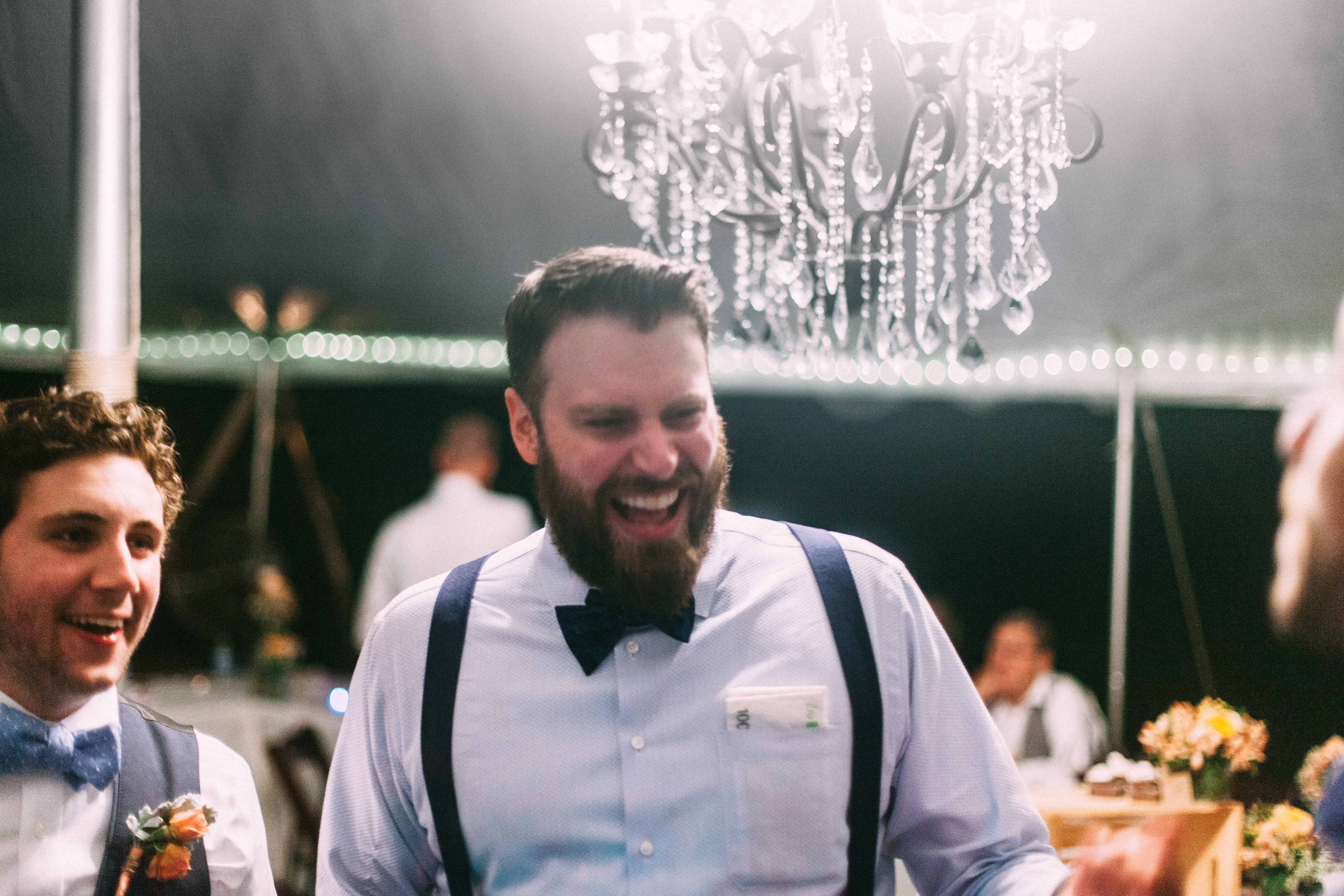 Bearded groom laughing heartily beside a groomsman under a crystal chandelier at a wedding reception.