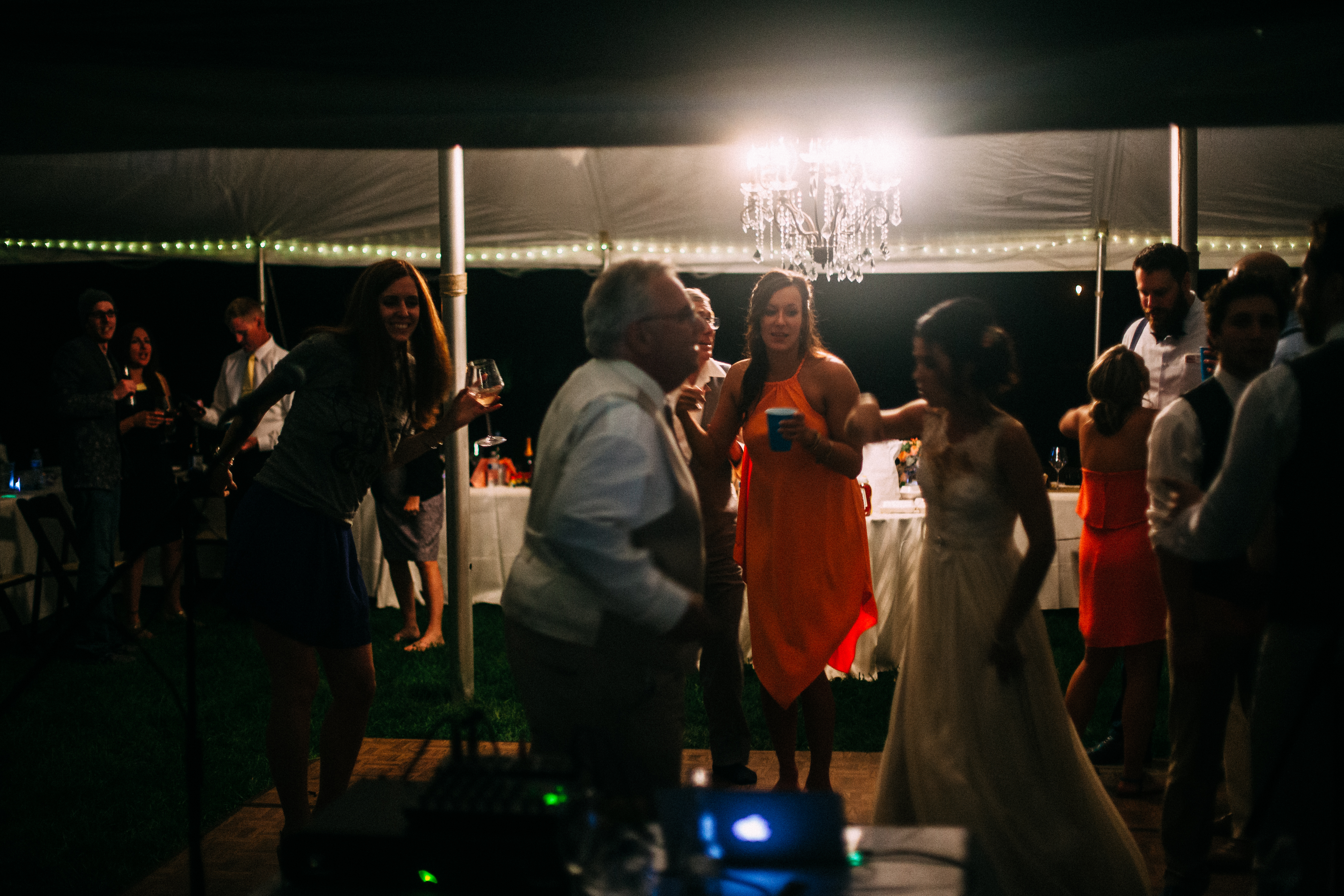 Bride and guests dancing outdoors on a wooden floor under an illuminated tent with crystal chandelier.