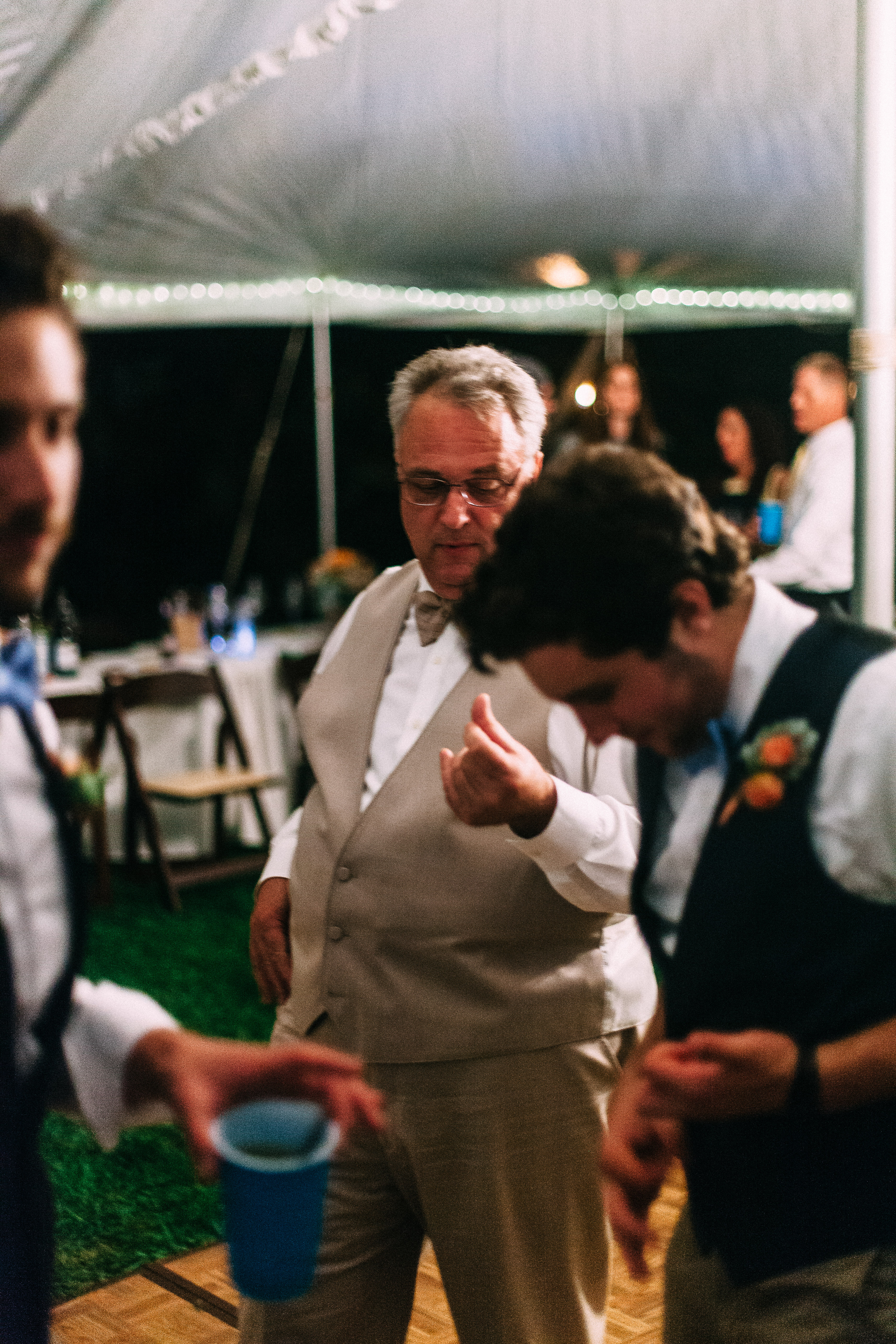 Older man in glasses and beige vest dancing with wedding guests on a wooden floor inside a tent.