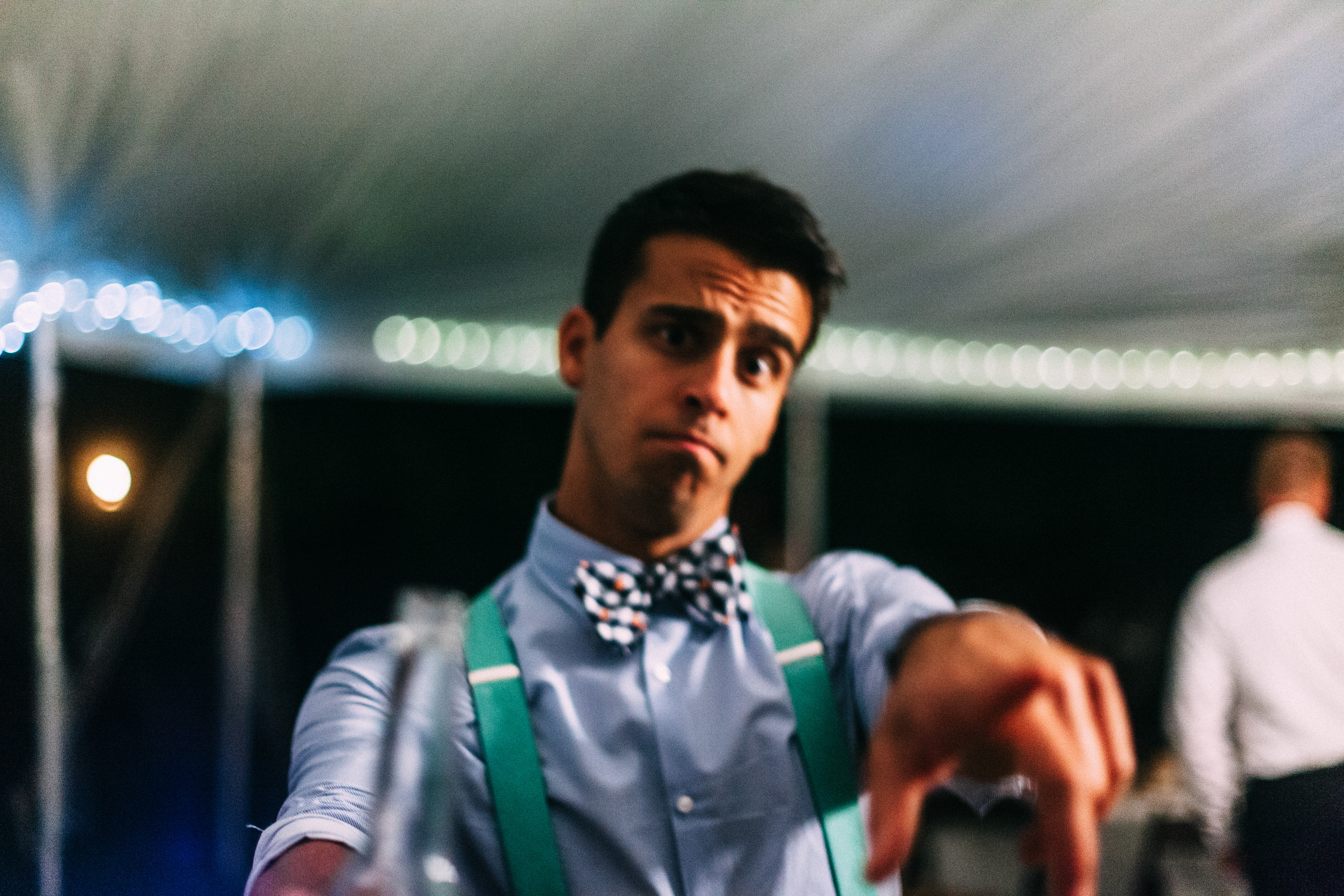 Groomsman in green suspenders and bow tie pointing playfully forward at a nighttime wedding reception.