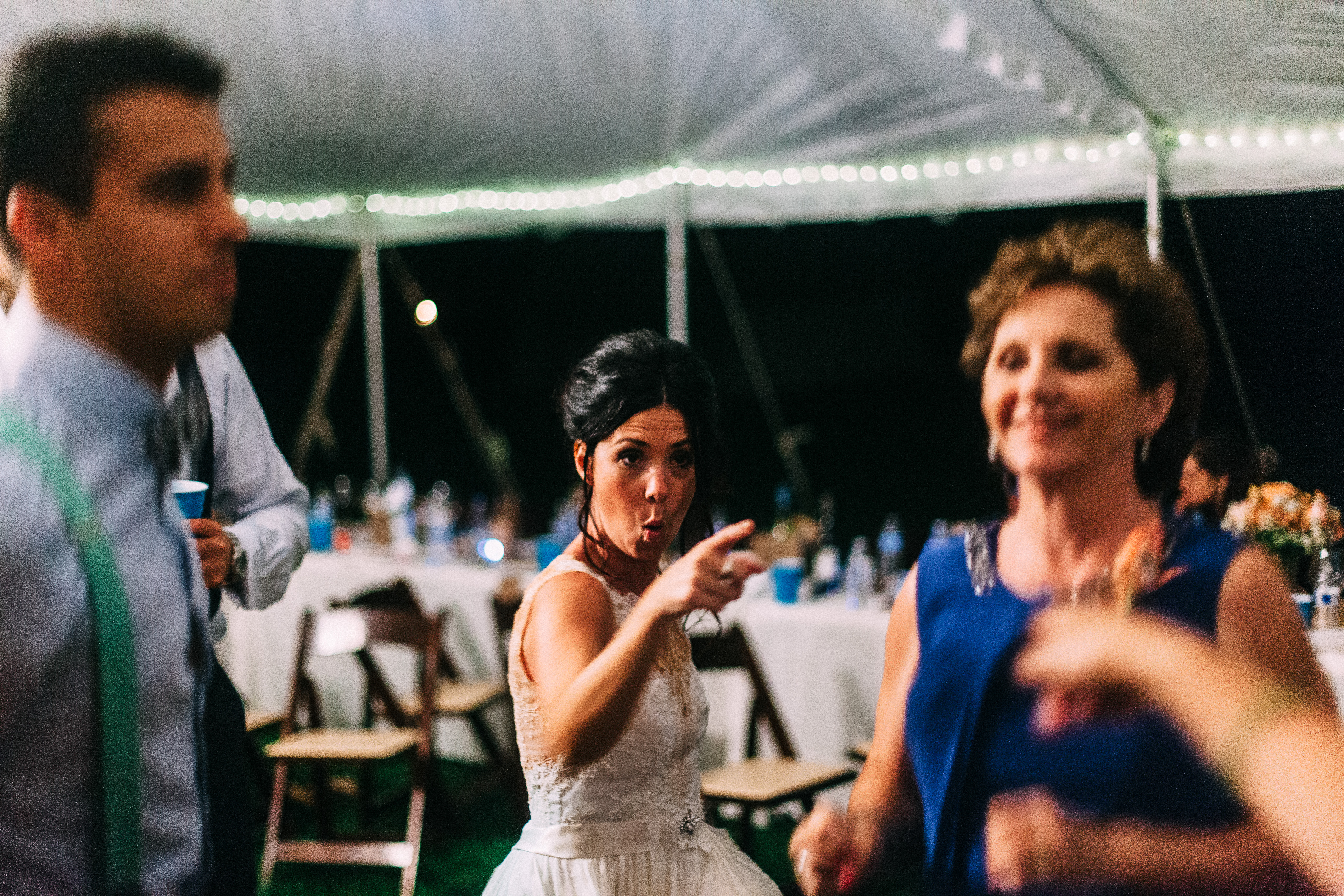 Bride in lace gown pointing enthusiastically on the dance floor next to guests in a reception tent.