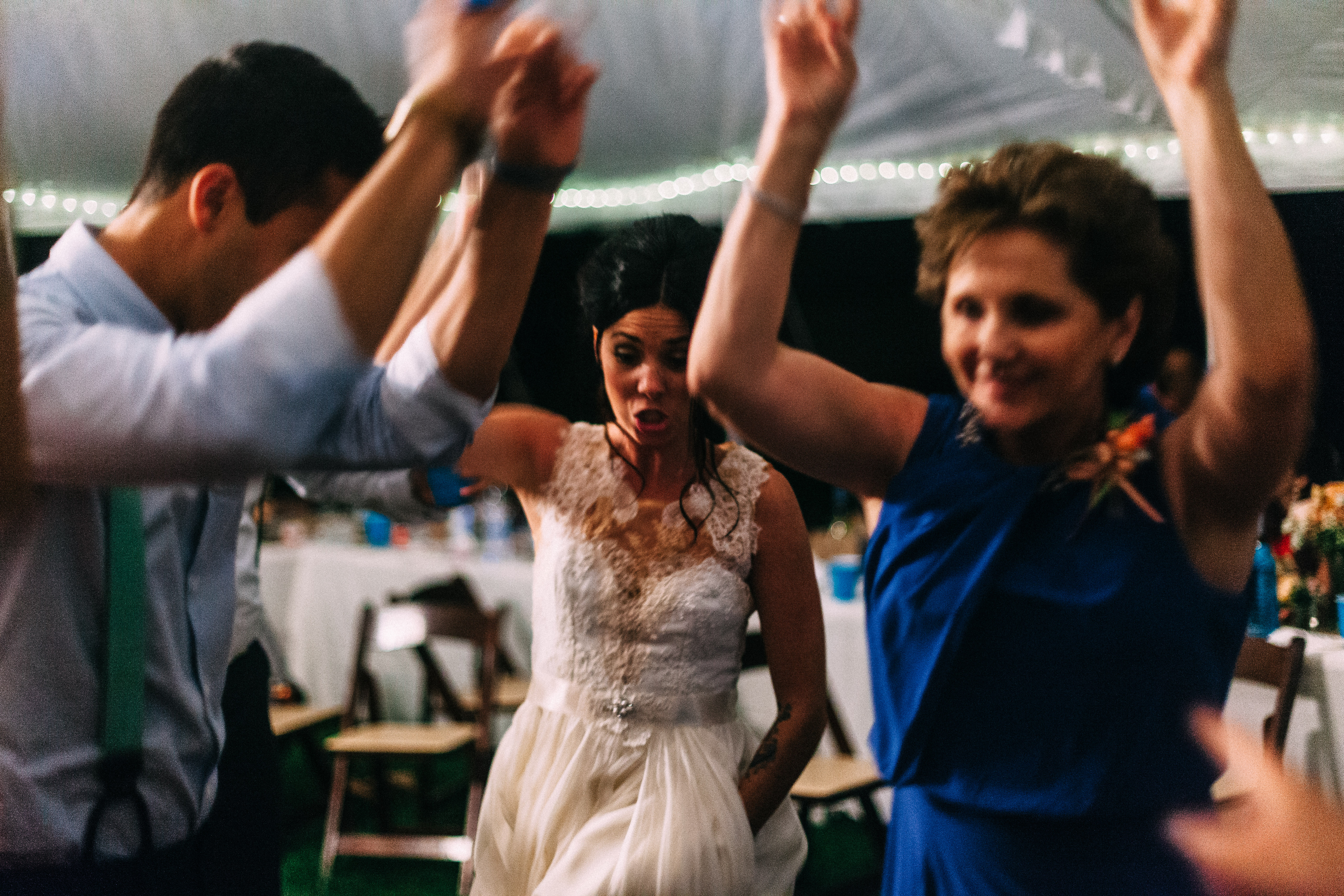 The bride and wedding guests dancing excitedly under an illuminated reception tent with hands raised in the air.