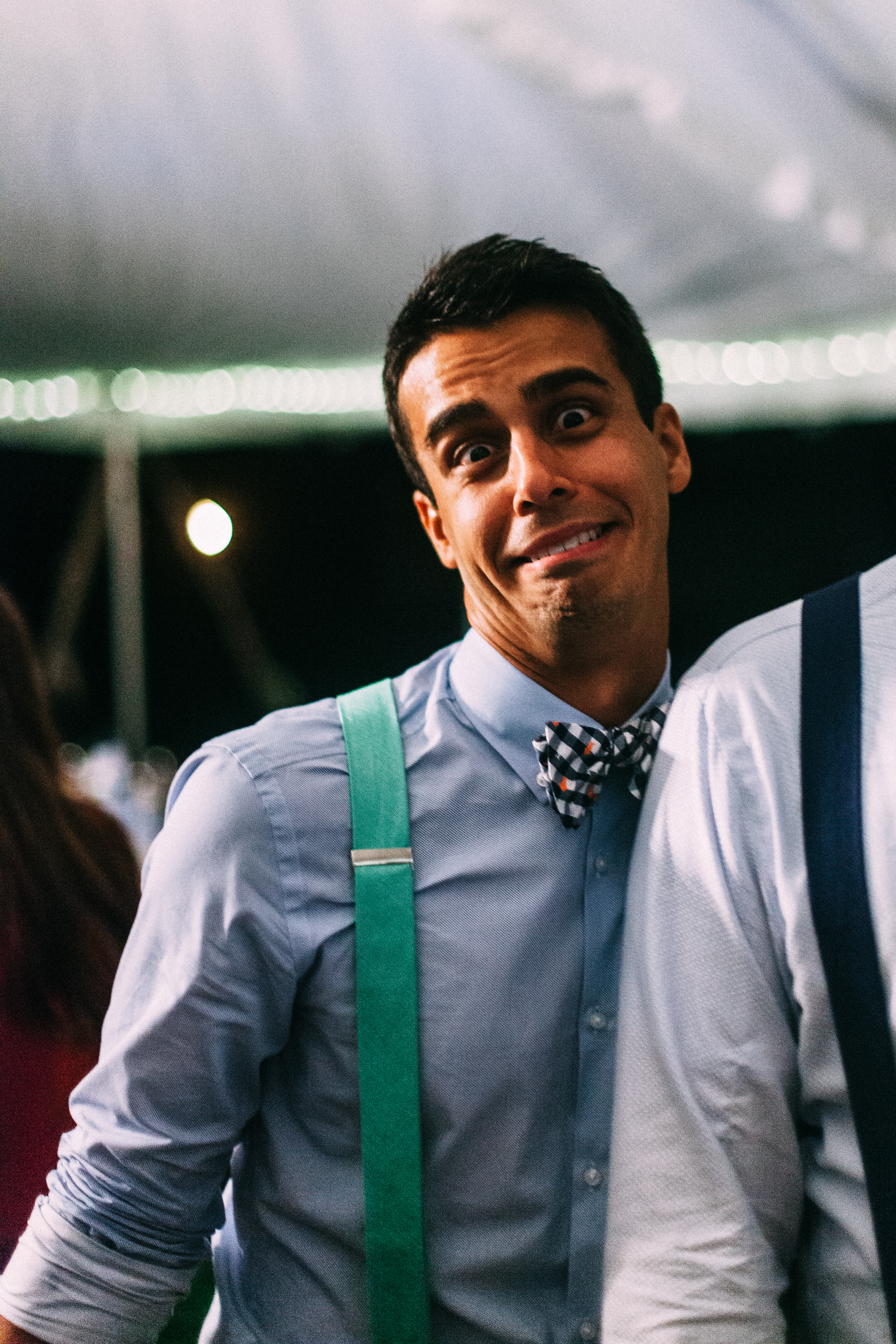 Groomsman in a bow tie and bright green suspenders making a humorous grimace at a wedding reception.