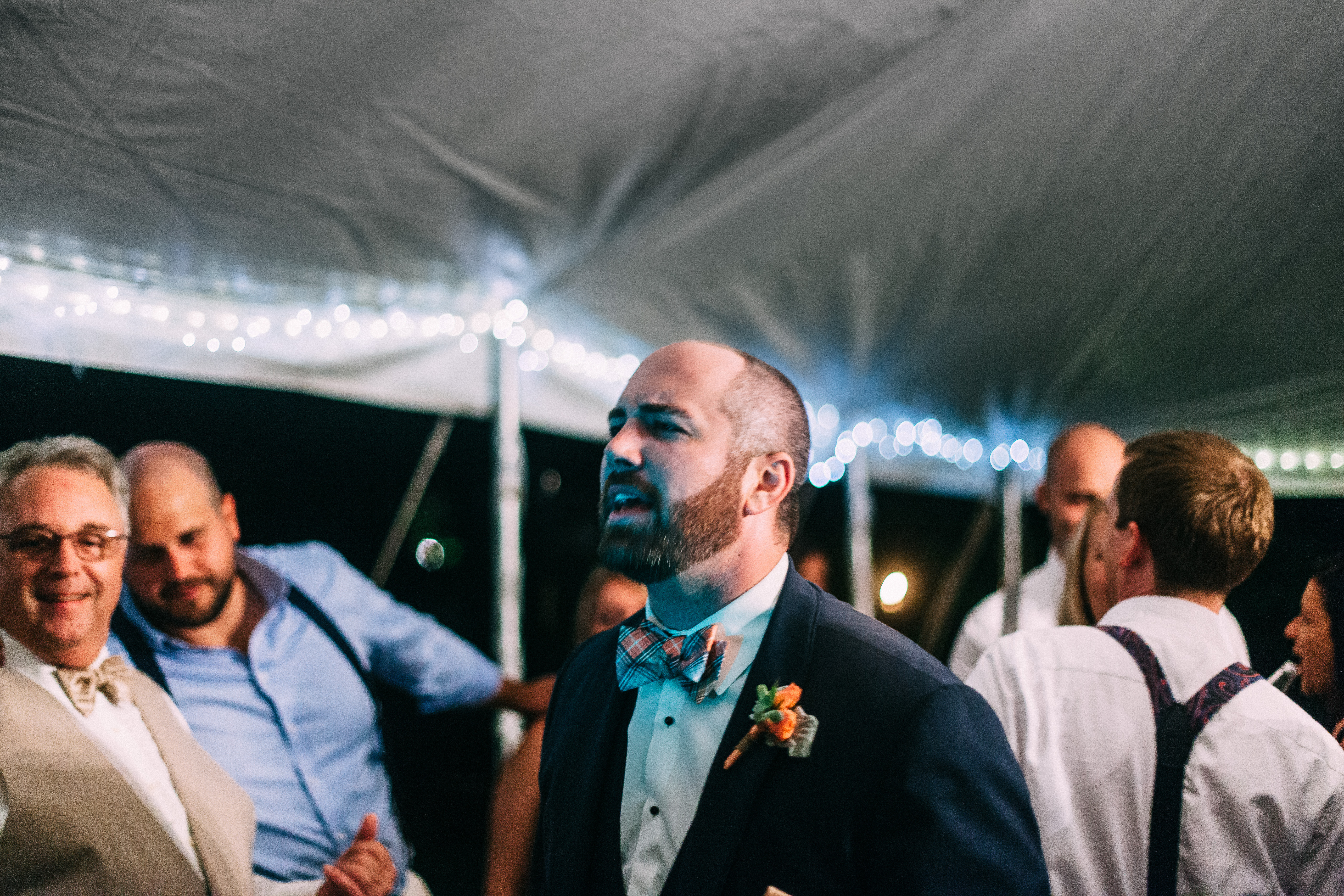 Groom singing along with smiling guests at a nighttime outdoor wedding reception under tent lighting.