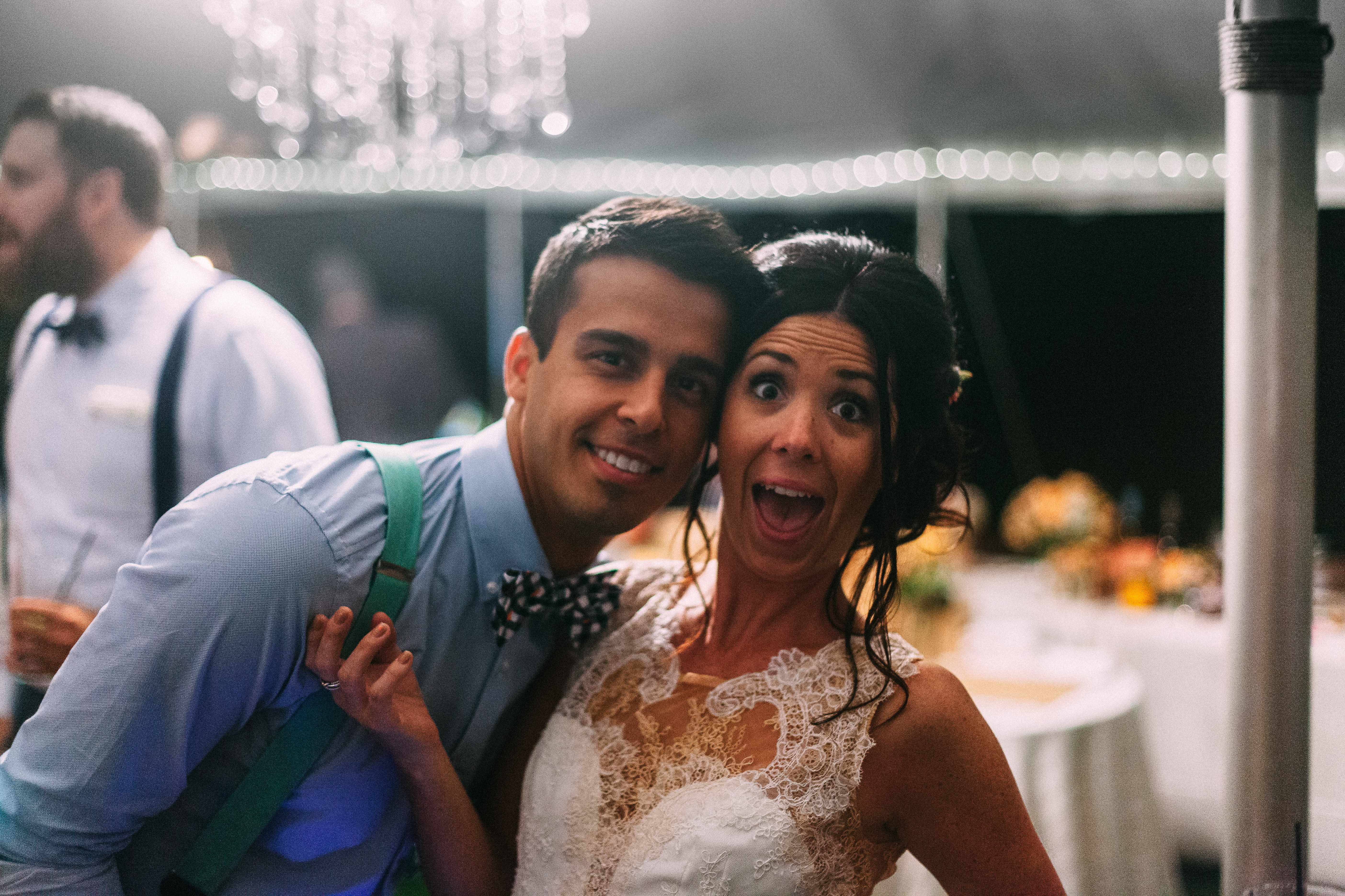 Bride with wide-eyed expression posing closely with a groomsman in green suspenders at wedding party.