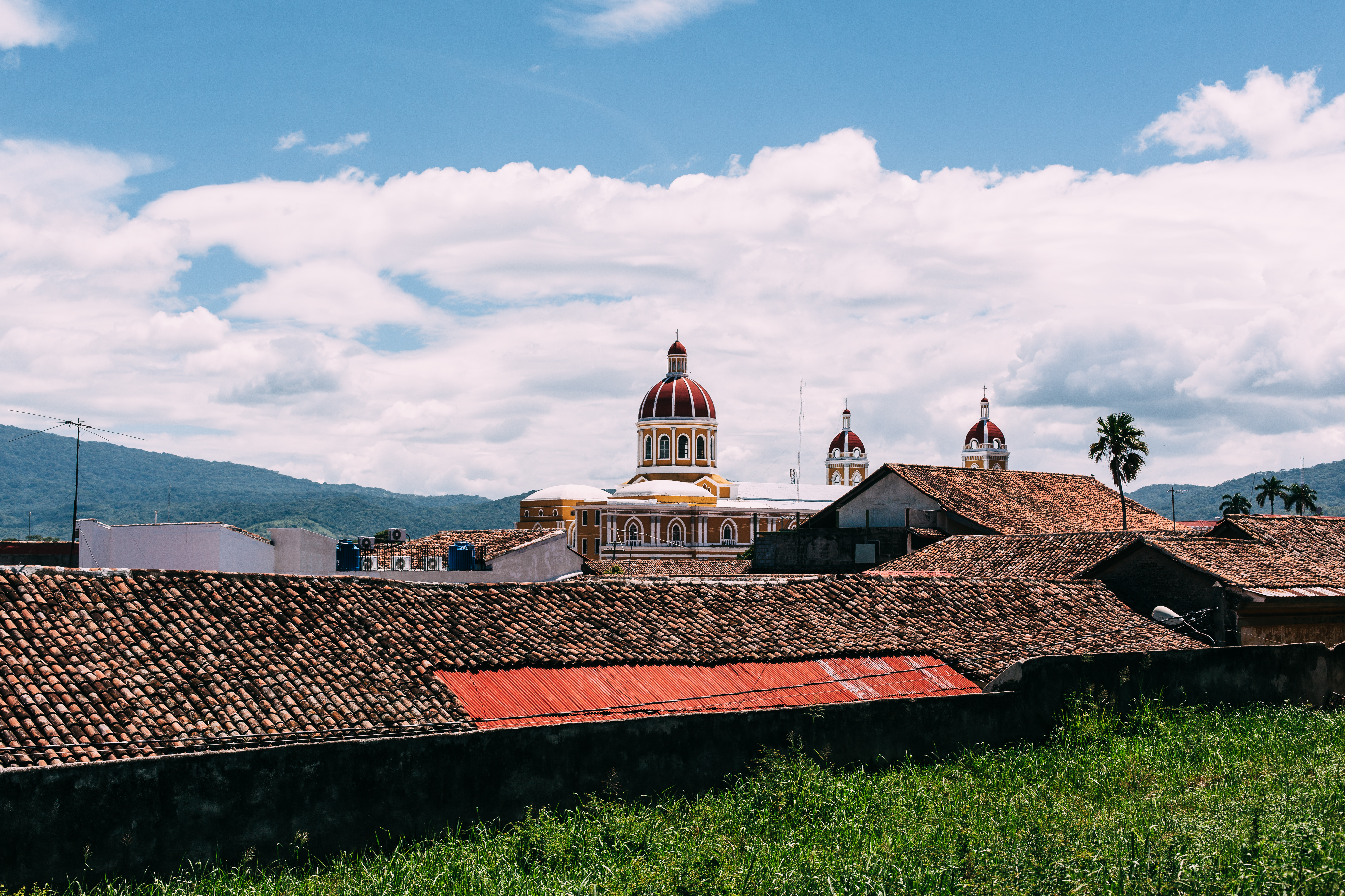 View over terracotta roofs toward yellow and red domes of Granada Cathedral against rolling green hills.