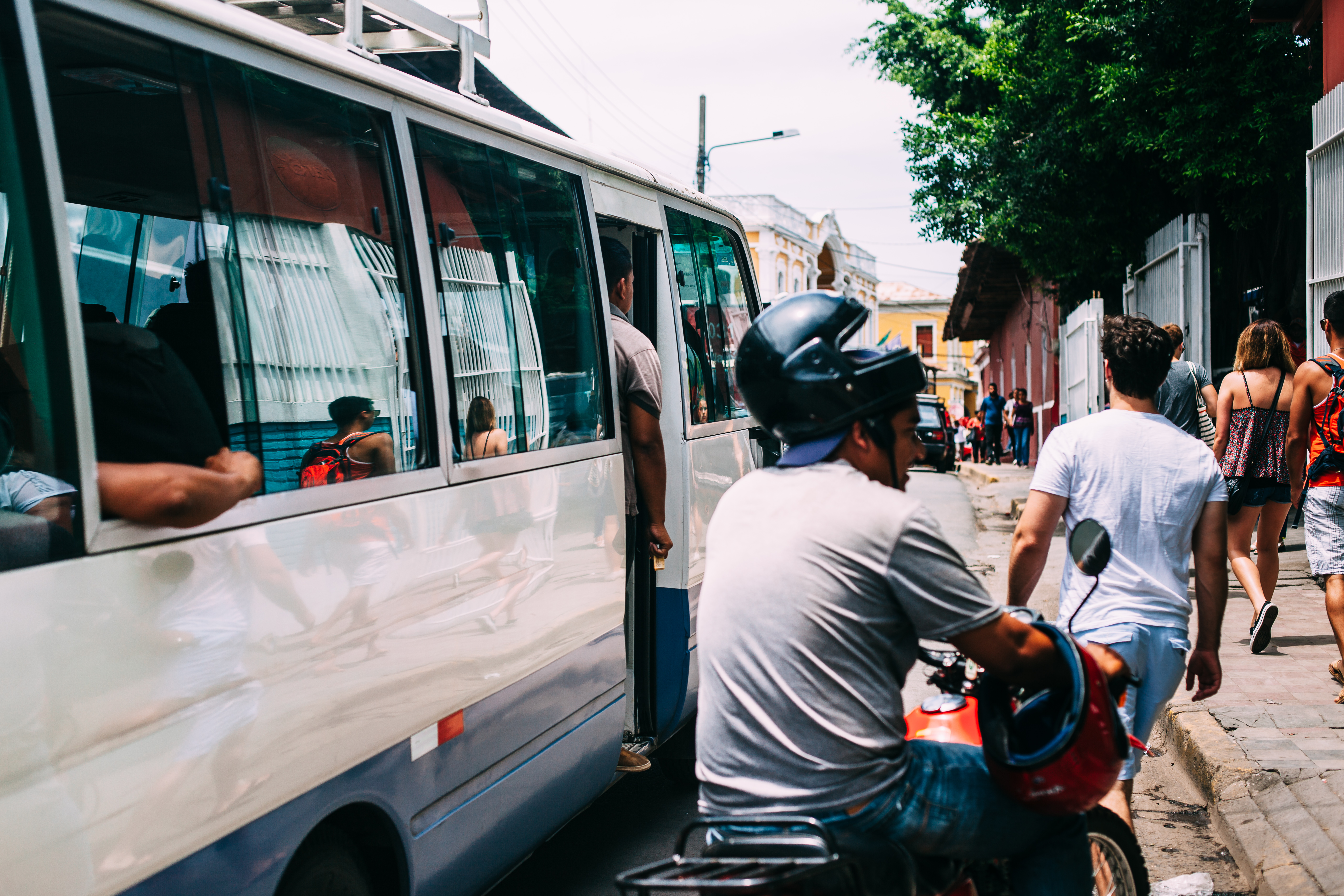 Minibus navigating a narrow street beside a motorcyclist and pedestrians in Granada, Nicaragua.