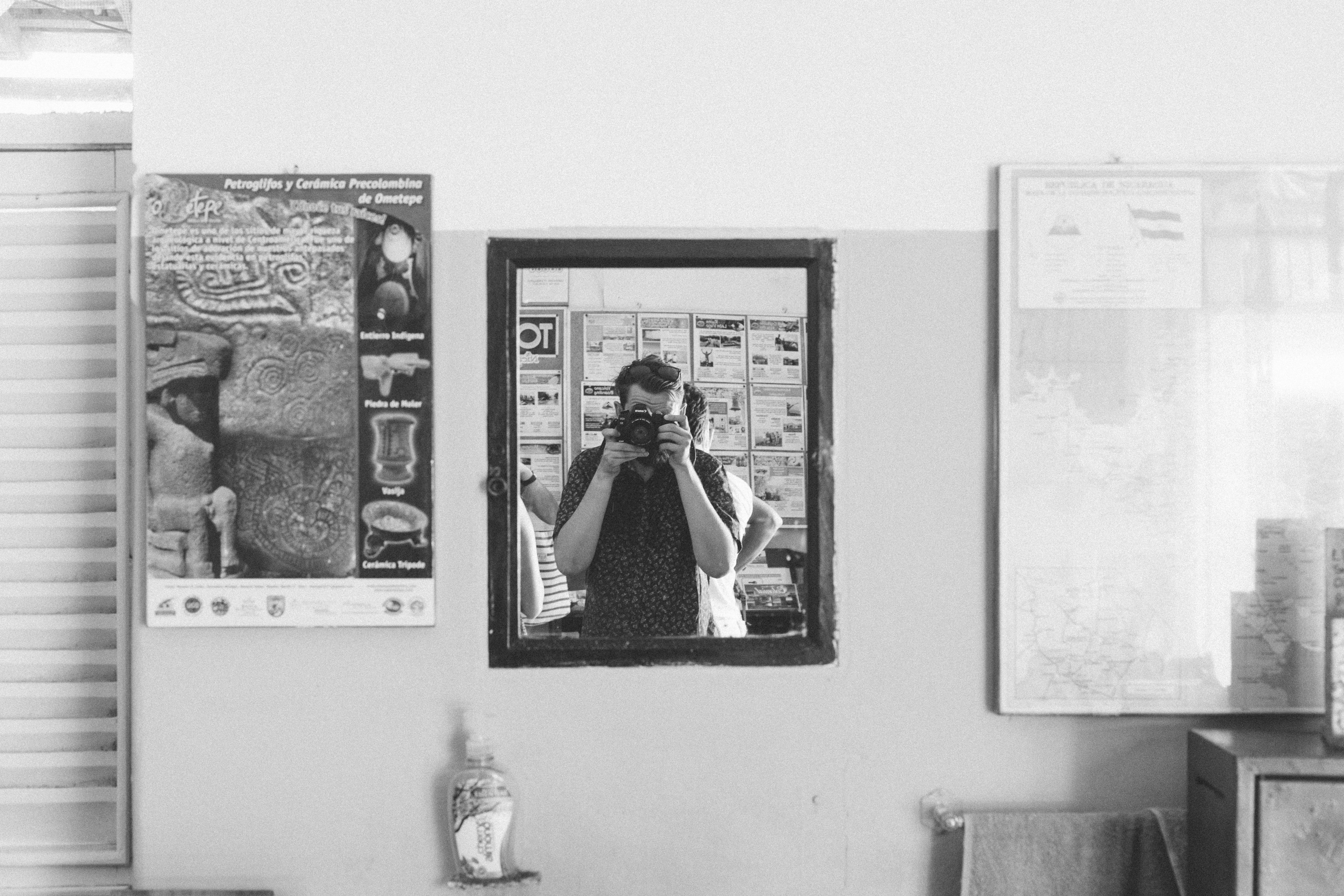 Mirror reflection showing a photographer holding a camera to his eye beside posters on a bathroom wall.