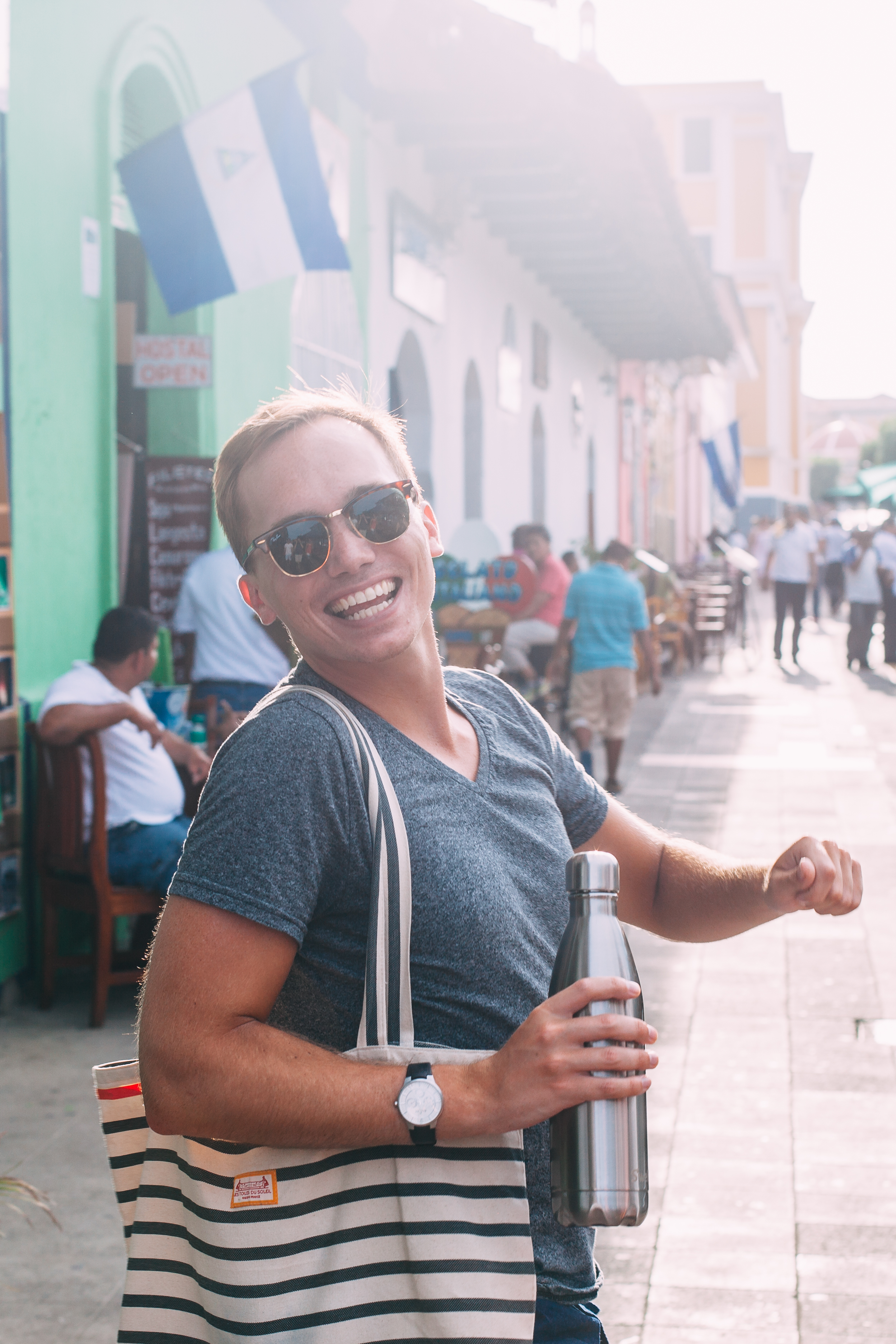 A smiling man wearing sunglasses turning towards the camera with a stainless steel water bottle in hand and a striped canvas tote bag on his shoulder on a sunlit street in Granada, Nicaragua, shot with a Canon 5D Mark II.