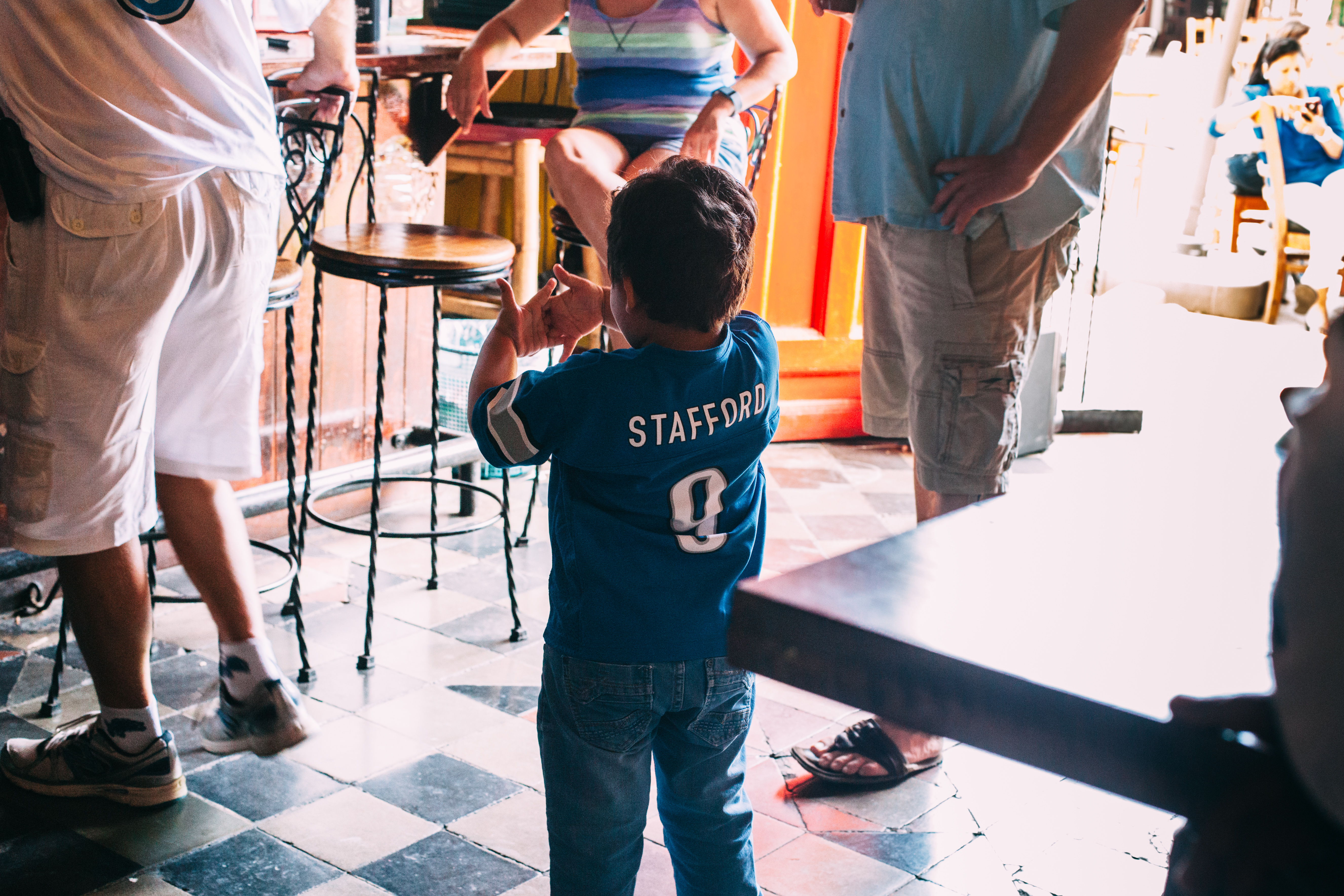 Back view of a young boy in a blue Detroit Lions Matthew Stafford #9 jersey and jeans clapping his hands in a restaurant with checkered tile floors in Granada, Nicaragua, shot with a Canon 5D Mark II.