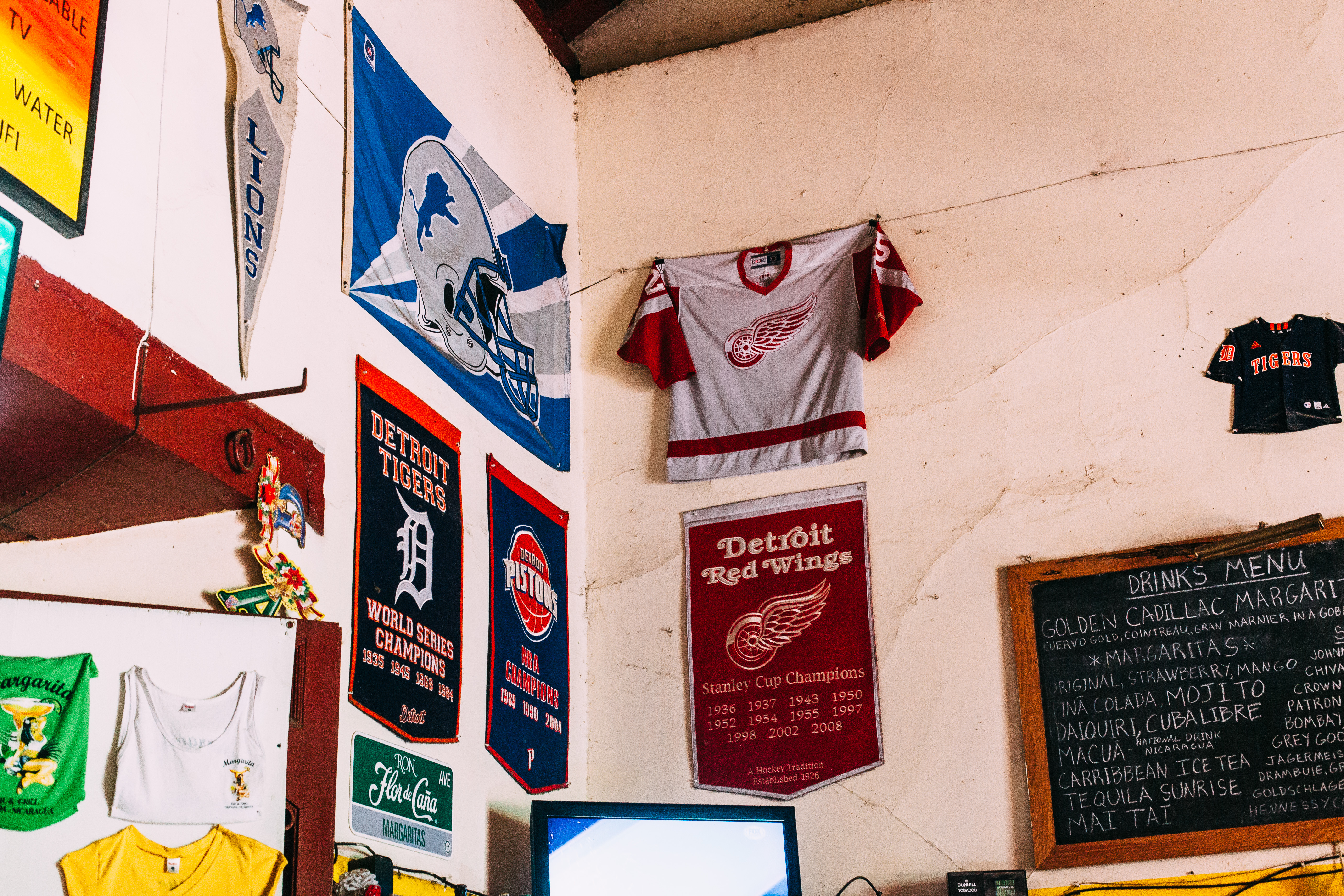 A wall decorated with Detroit sports memorabilia, including a Detroit Lions flag and pennant, Detroit Tigers championship banner, Detroit Red Wings jersey and banner, and a handwritten drinks chalkboard in Granada, Nicaragua, shot with a Canon 5D Mark II.