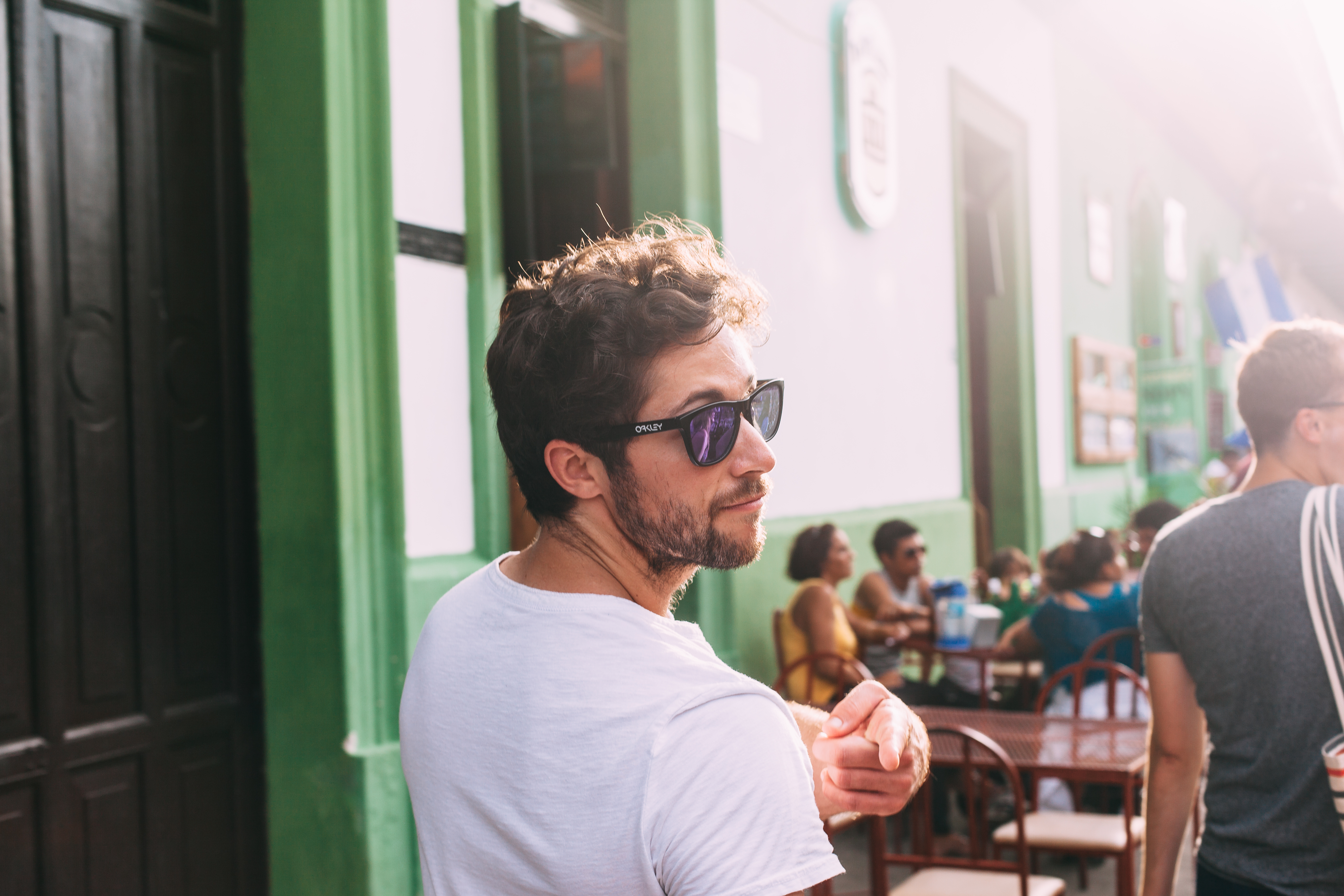 Profile of a bearded young man in sunglasses and a white t-shirt gesturing with his hand while walking past an outdoor café terrace in Granada, Nicaragua, shot with a Canon 5D Mark II.