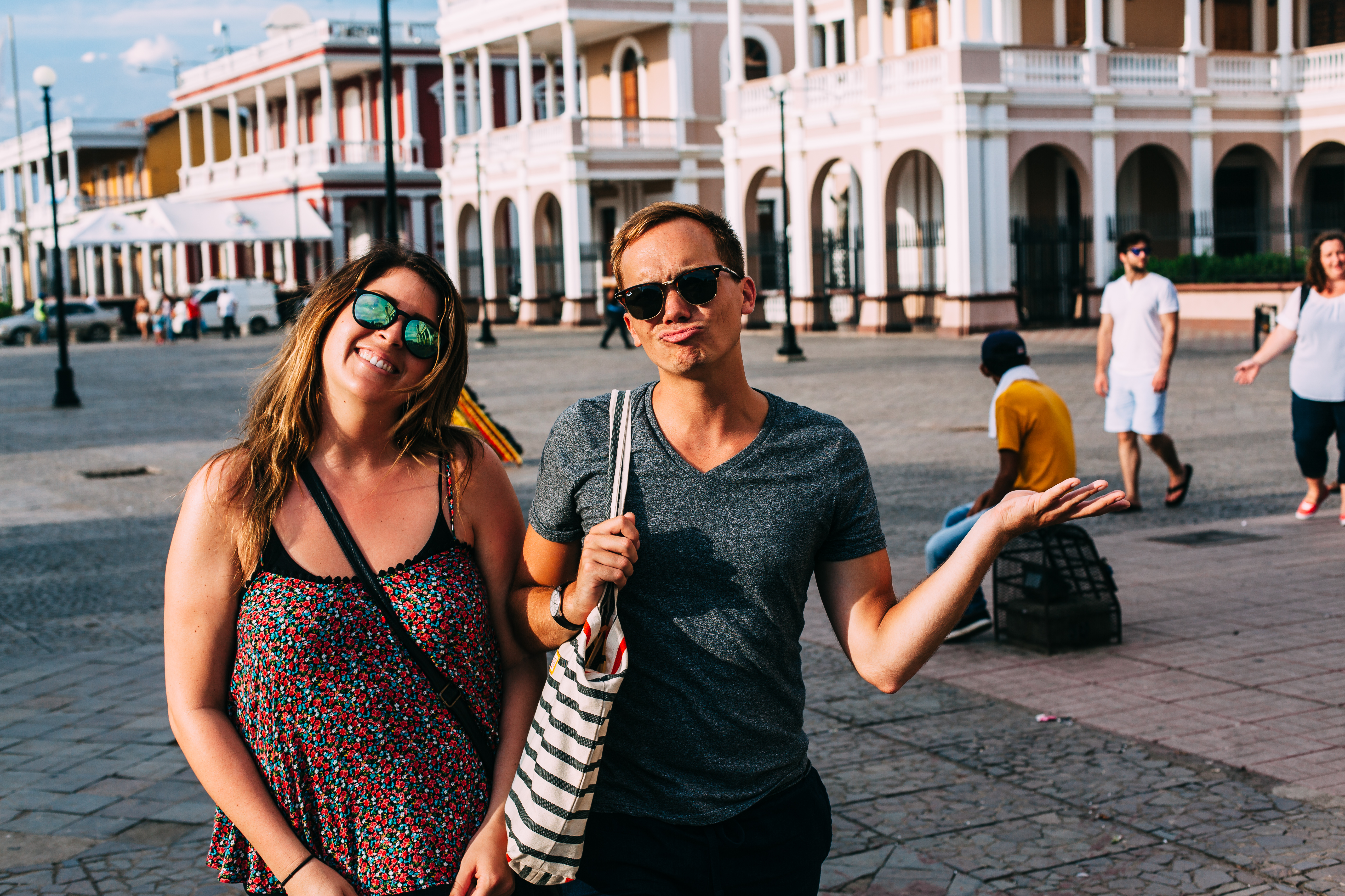 A smiling woman in a floral sundress and a man playfully shrugging with an outstretched hand standing together in the sunlit town square plaza of Granada, Nicaragua, shot with a Canon 5D Mark II.