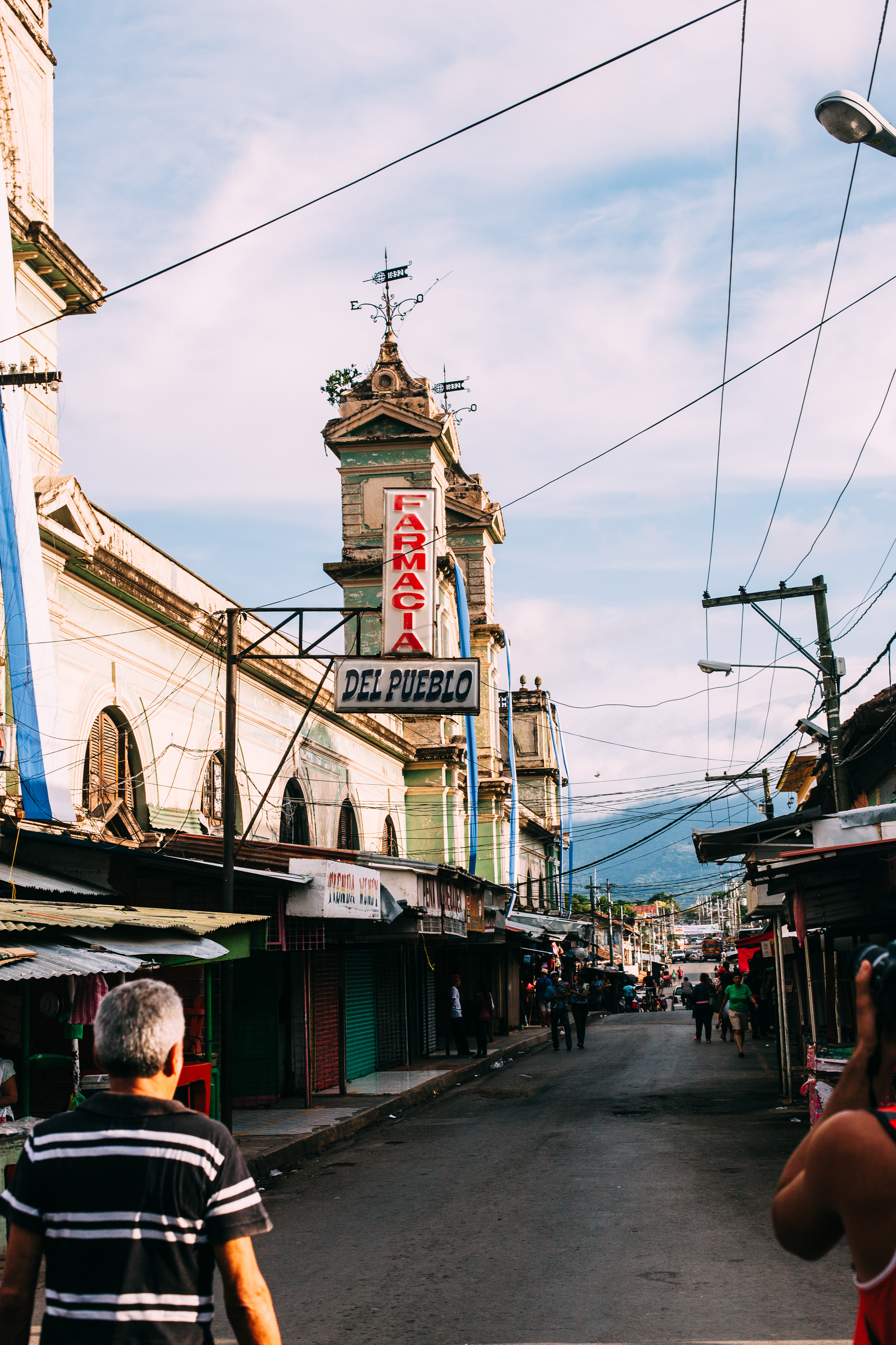 Market street in Granada, Nicaragua with shops, pedestrians, and a vertical 'FARMACIA DEL PUEBLO' sign.