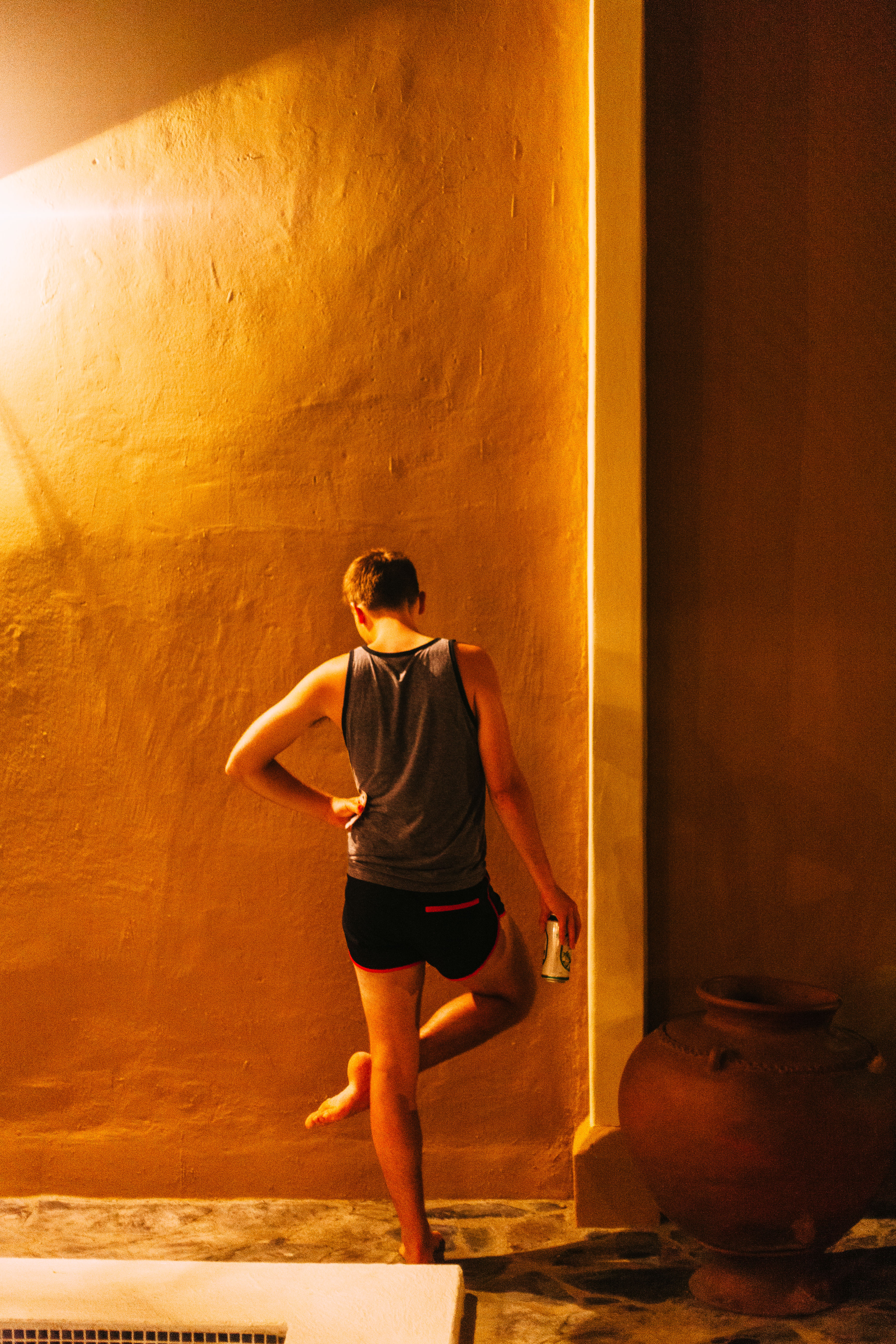 Young man in a grey tank top leaning against a warm amber-lit stucco wall beside a ceramic urn by a pool.