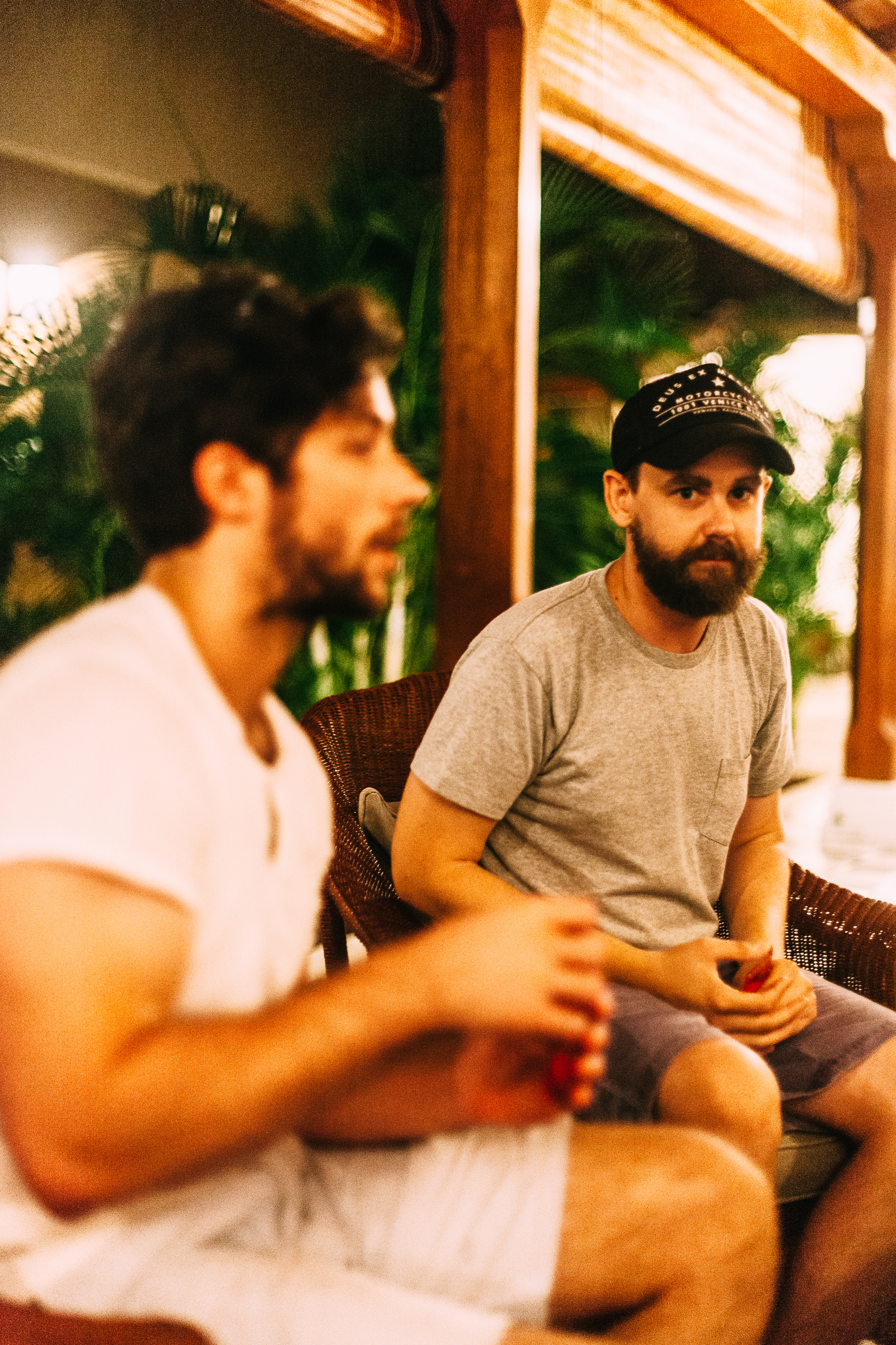 Two young men in casual attire sitting in wicker chairs on an outdoor patio in warm evening light.