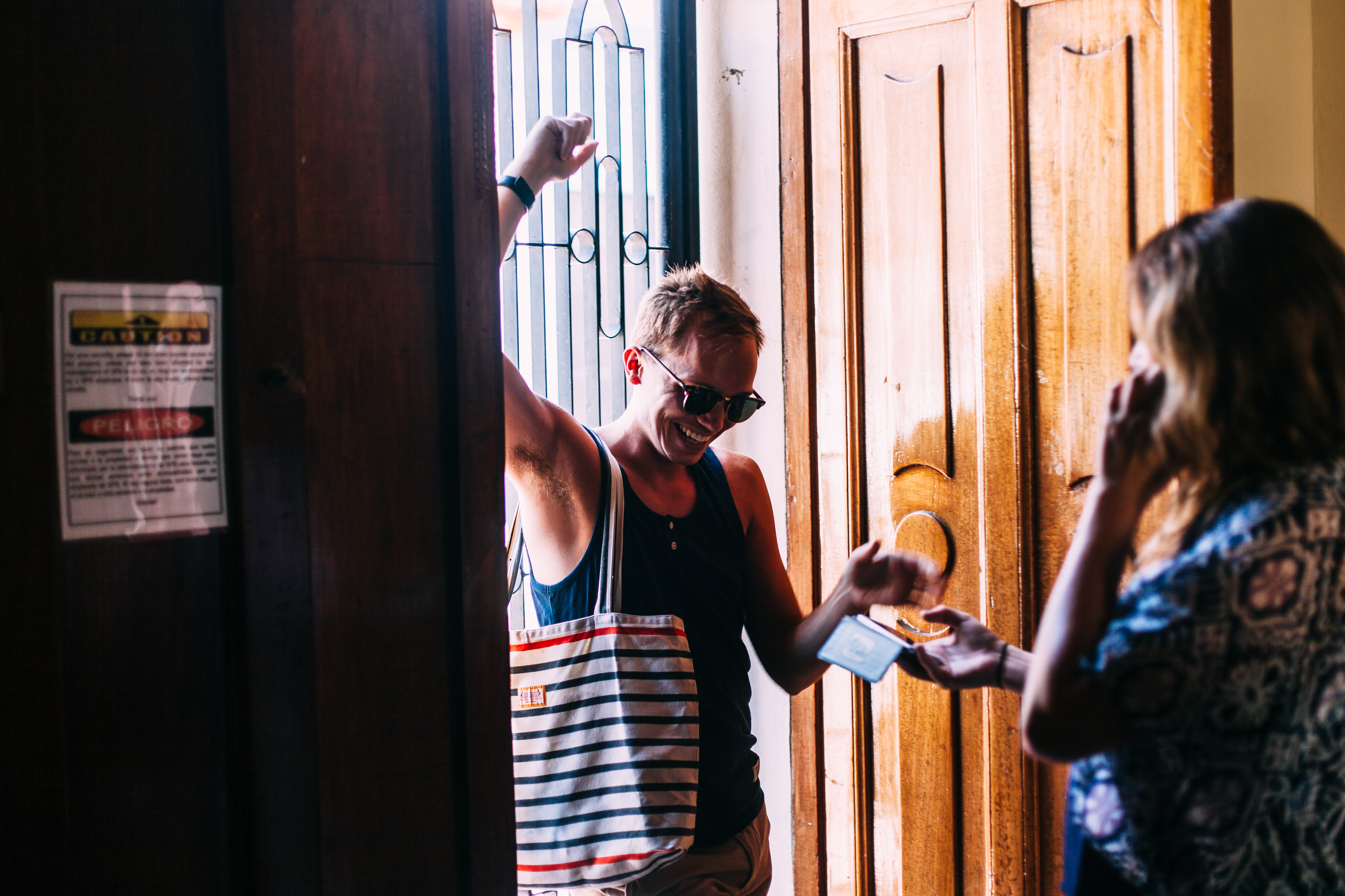 Smiling young man in sunglasses and striped tote bag raising his arm in excitement in a wooden doorway.