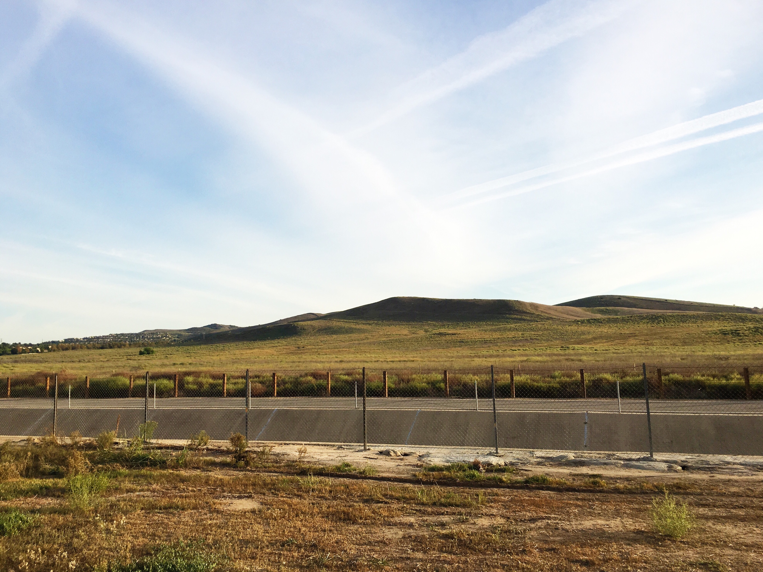 Paved roadway along a chain-link fence stretching across dry golden fields toward rolling brown hills.