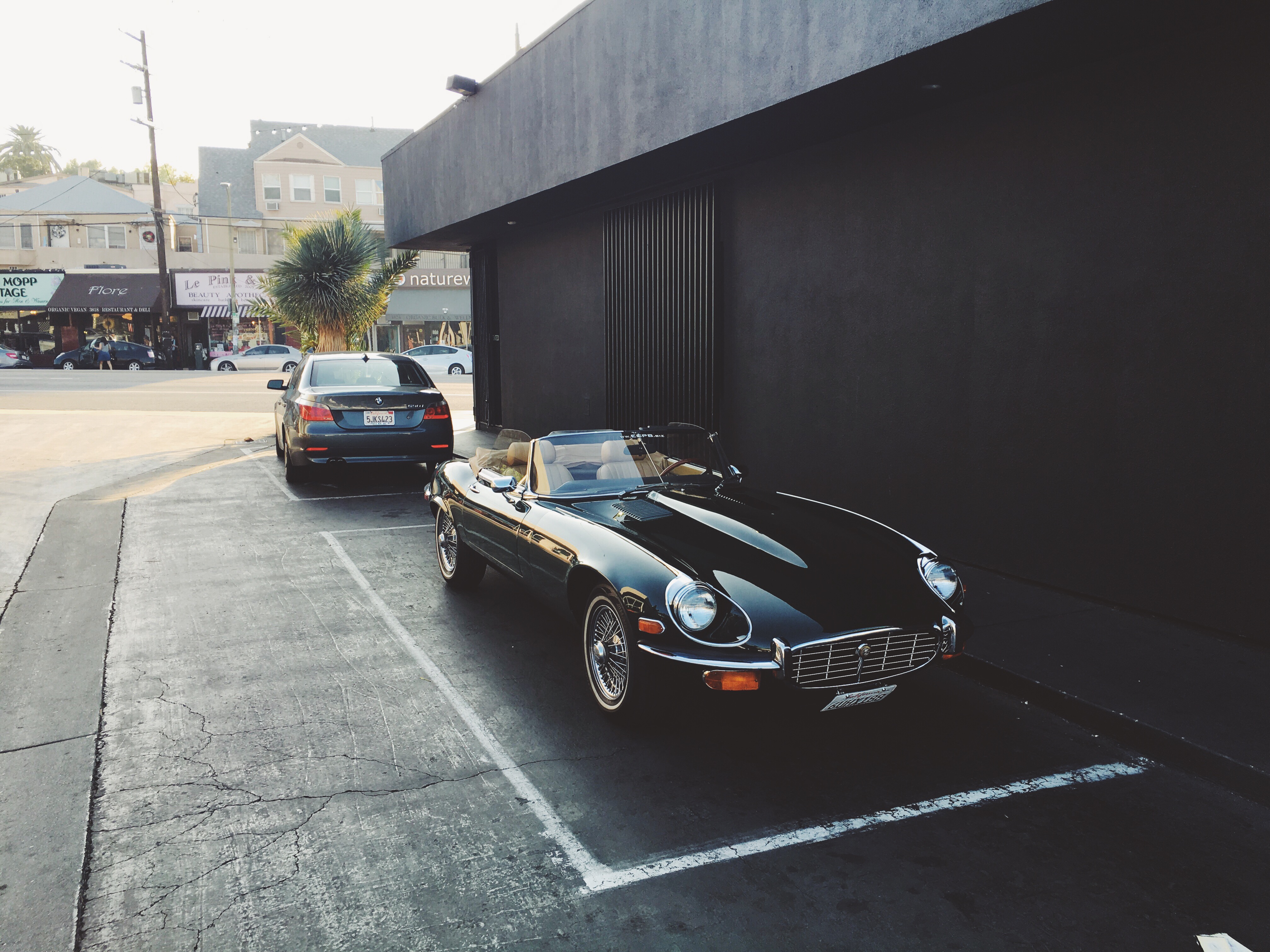 A classic black Jaguar E-Type roadster convertible with wire wheels and tan leather interior parked in an asphalt parking space in front of a modern matte black building on a sunny California street, shot with an iPhone 6s.