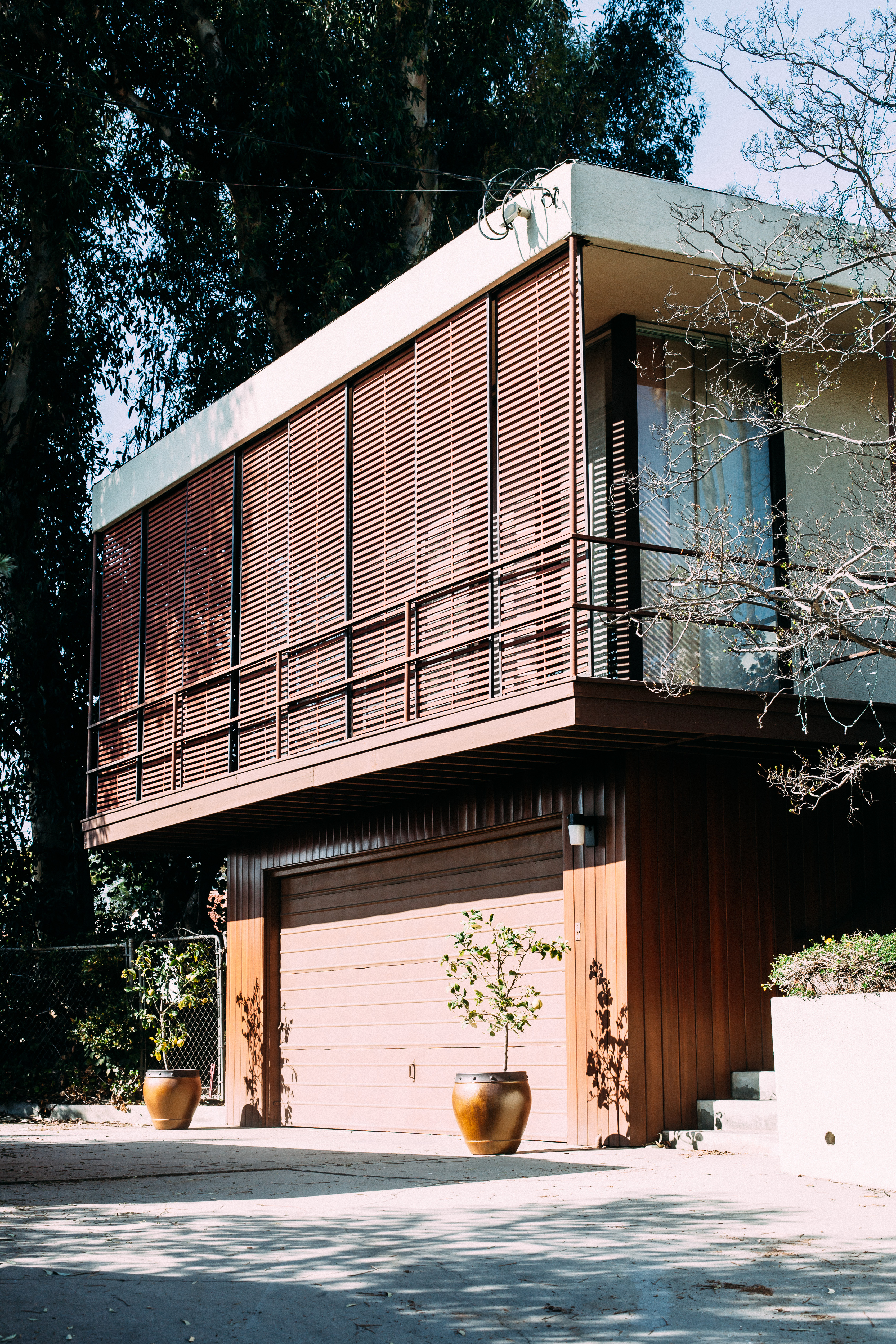 Mid-century two-story home with wood louvers on upper balcony over a garage with potted driveway plants.