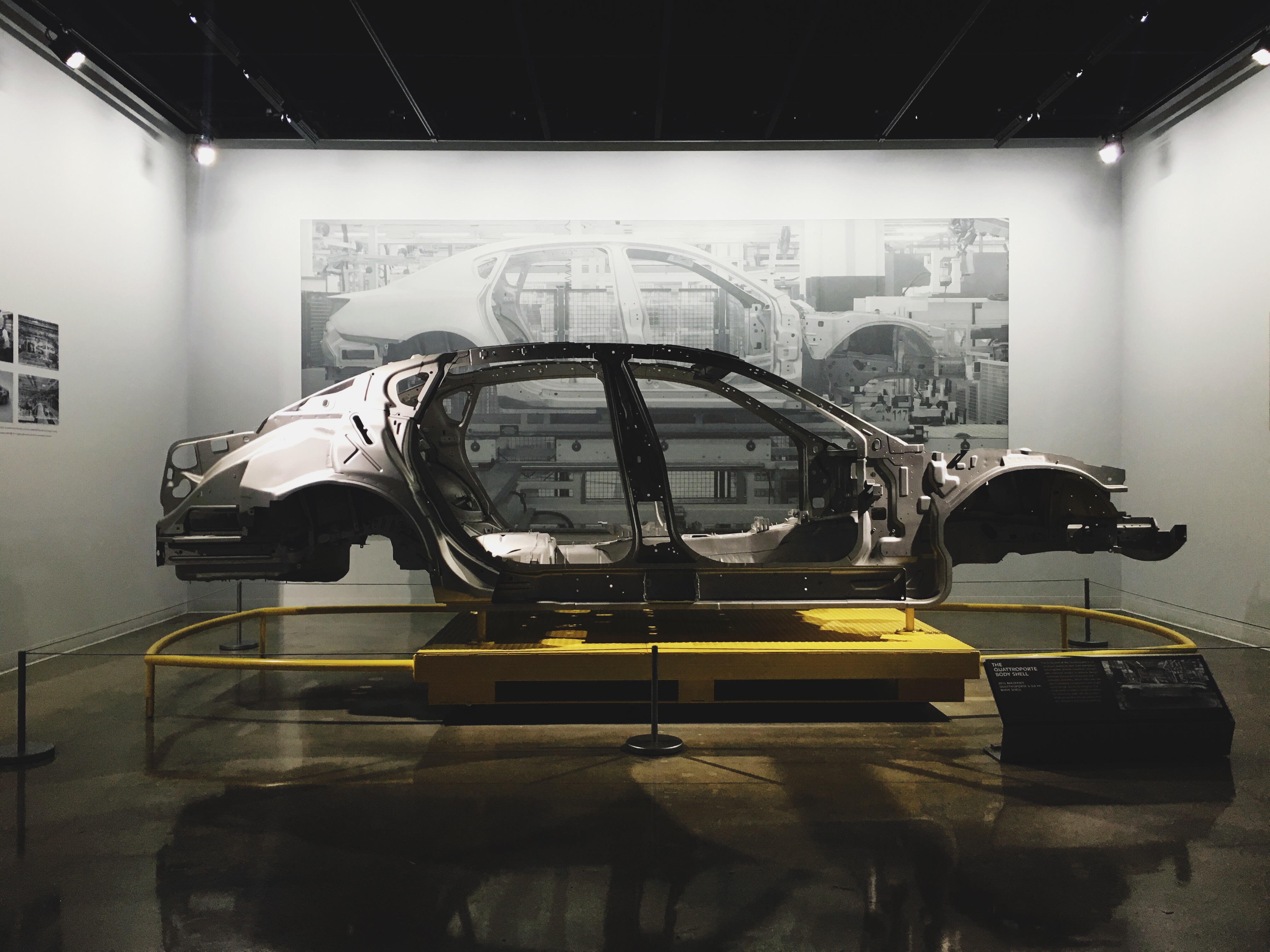 Bare automotive unibody frame mounted on a yellow exhibition stand against a factory assembly mural.