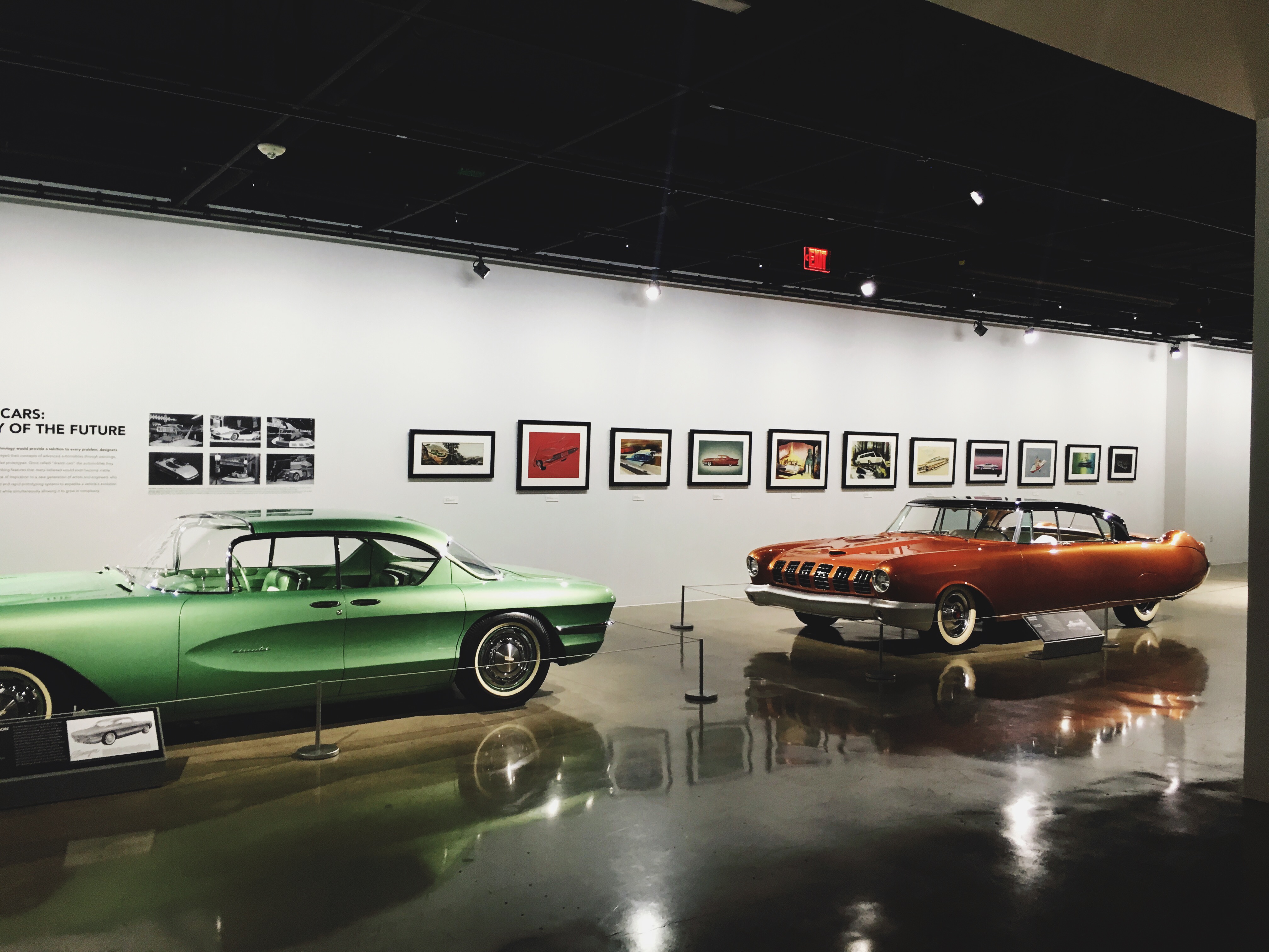 Mid-century concept cars in green and copper on a reflective museum floor beneath framed design art.