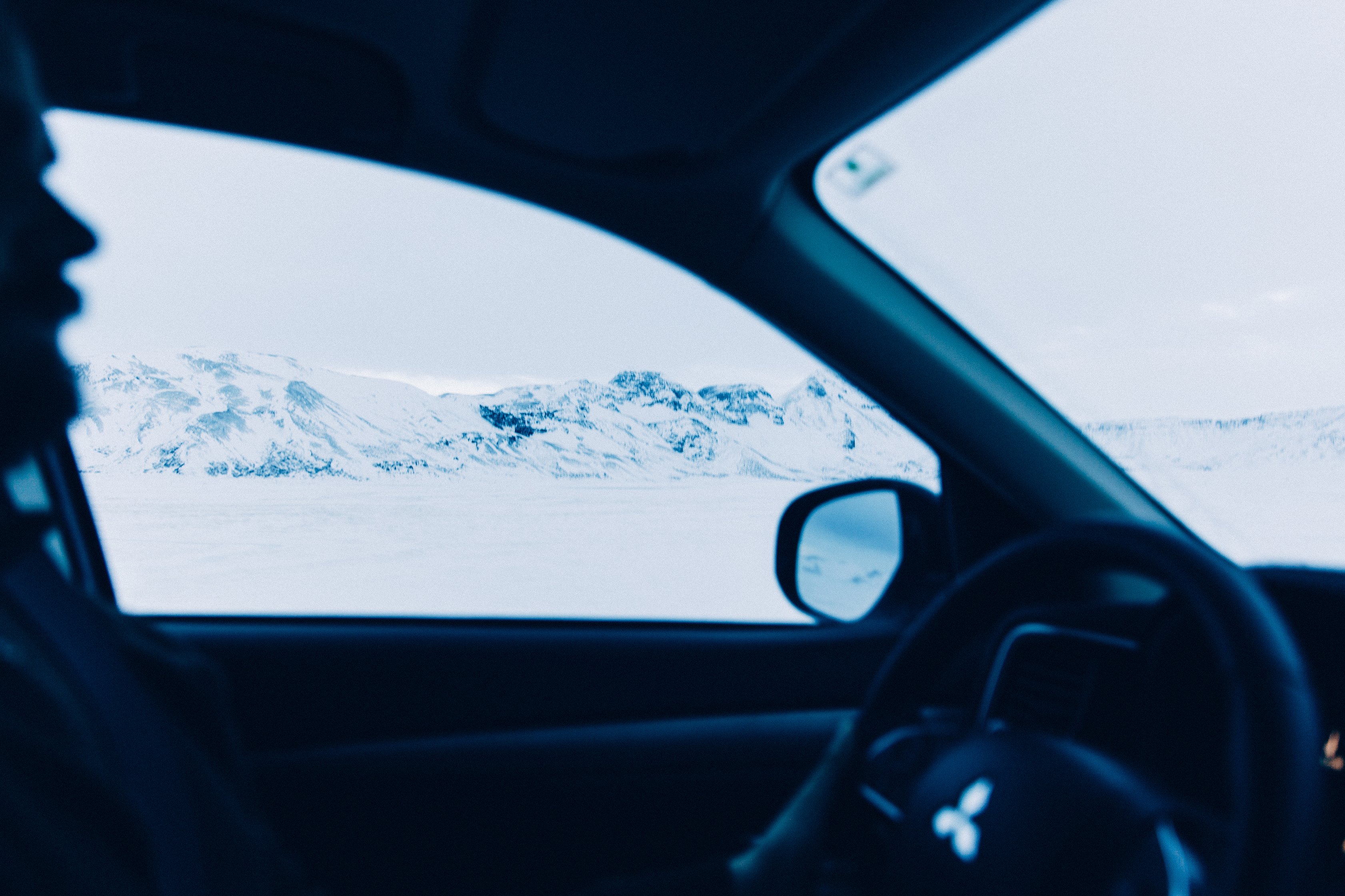 Driver silhouette looking out across a snow-covered valley toward jagged snowy mountains in Iceland.