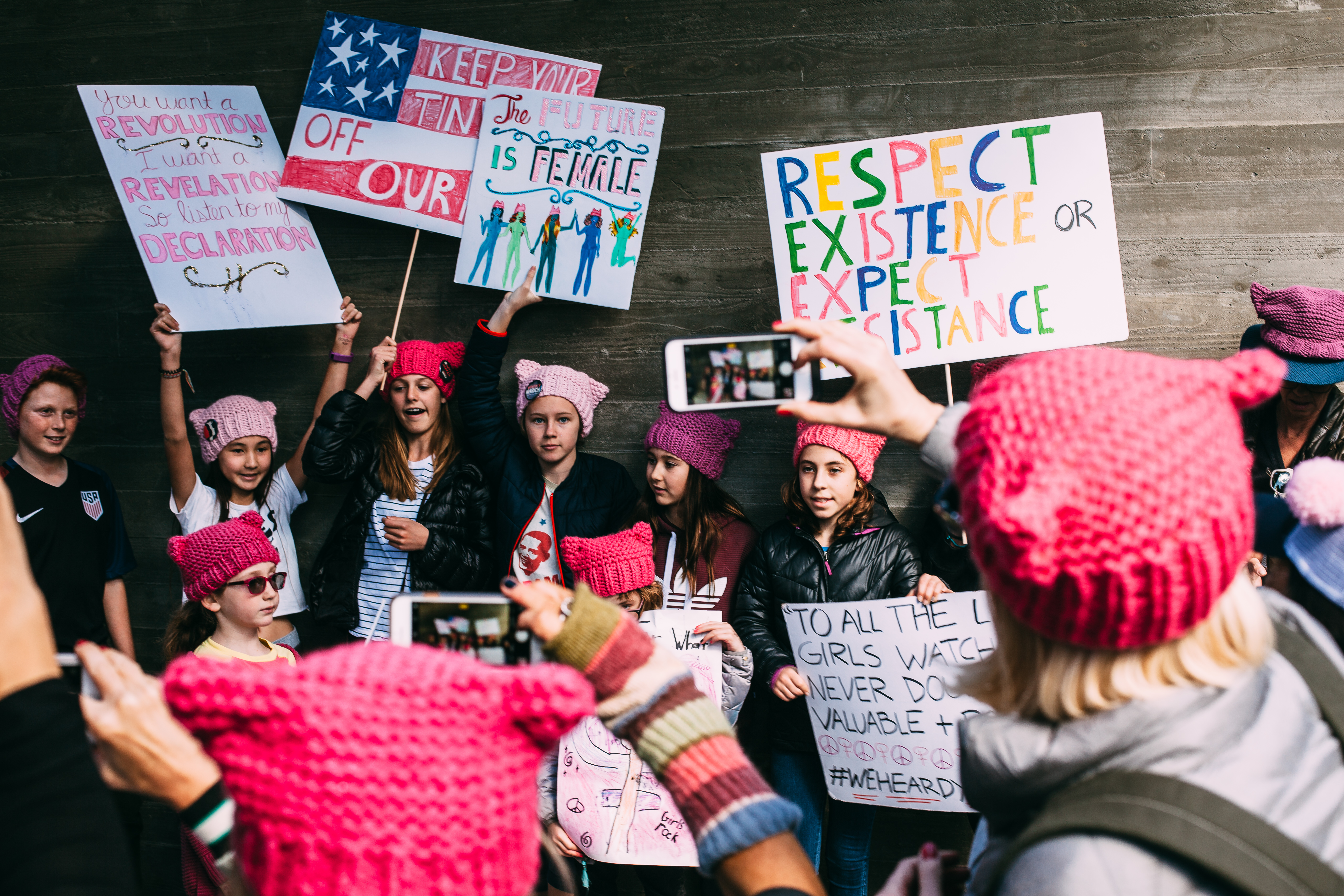 Young girls wearing pink knit pussyhats holding handmade protest signs while people take photos with smartphones at the Women's March in Los Angeles, shot with a Canon 5D Mark II.