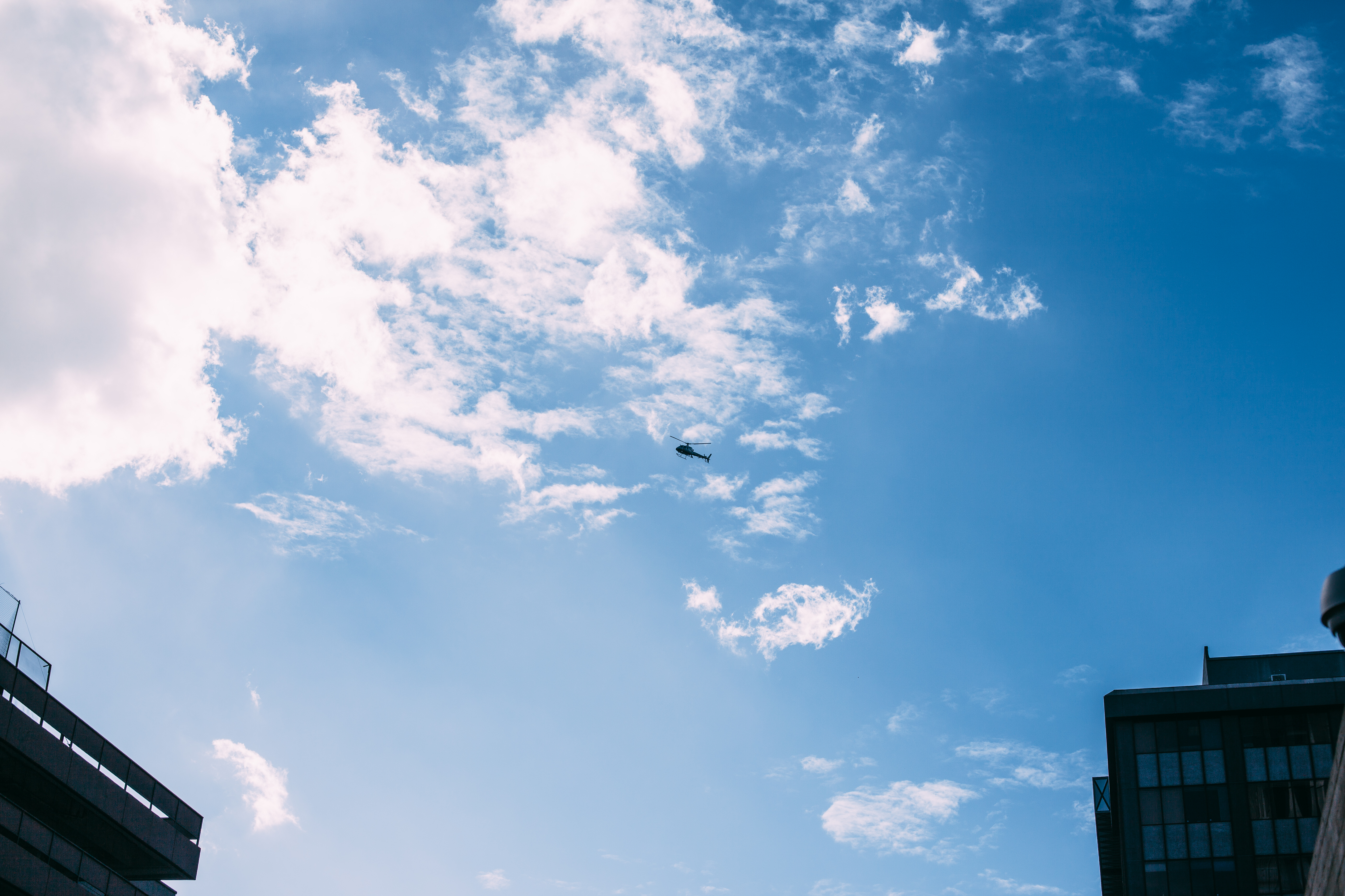 A news helicopter hovering high in a bright blue sky with wispy clouds between downtown Los Angeles buildings above the Women's March, shot with a Canon 5D Mark II.