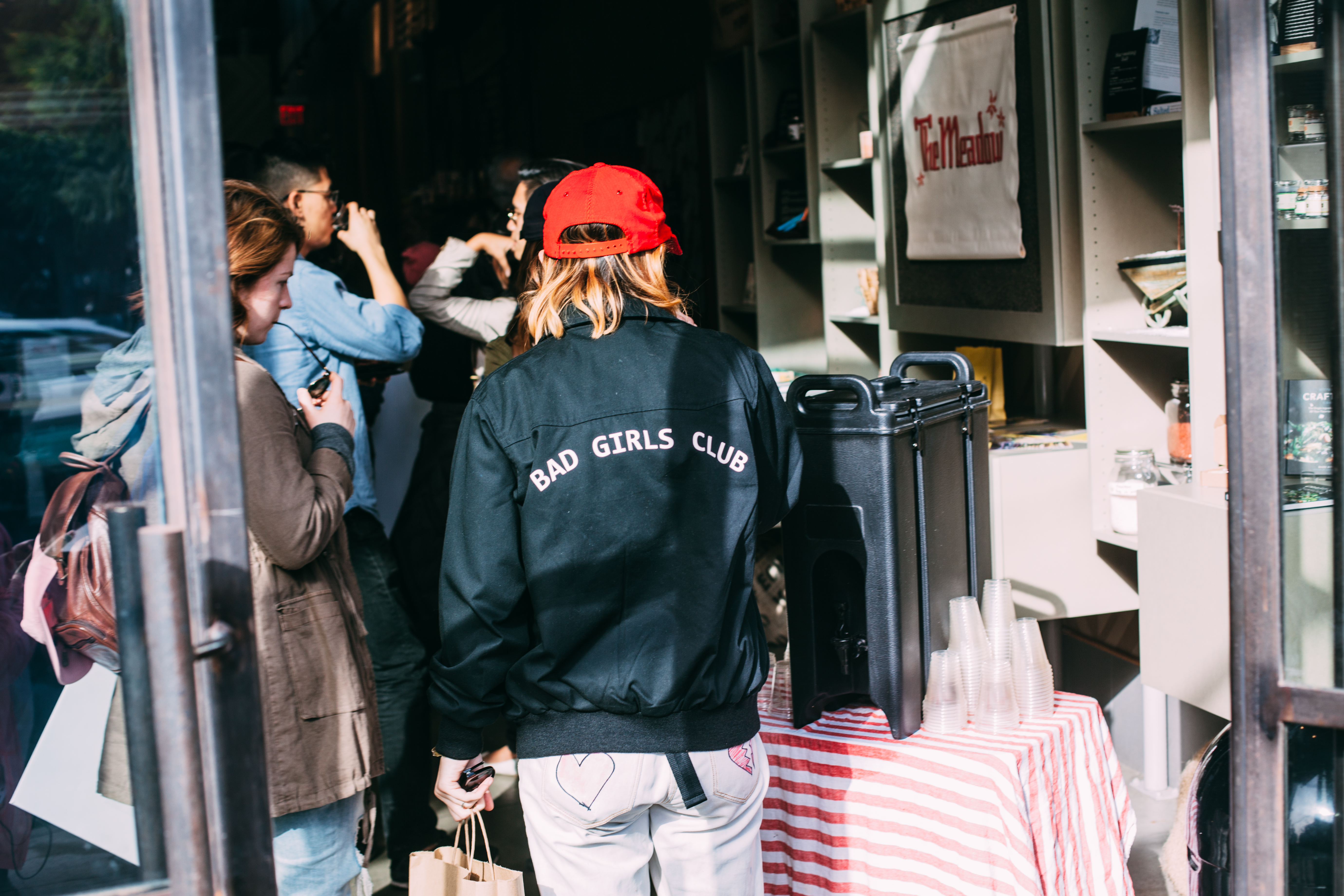 A woman wearing a red cap and a black jacket embroidered with 'BAD GIRLS CLUB' on the back standing by an open shop doorway at the Women's March in Los Angeles, shot with a Canon 5D Mark II.