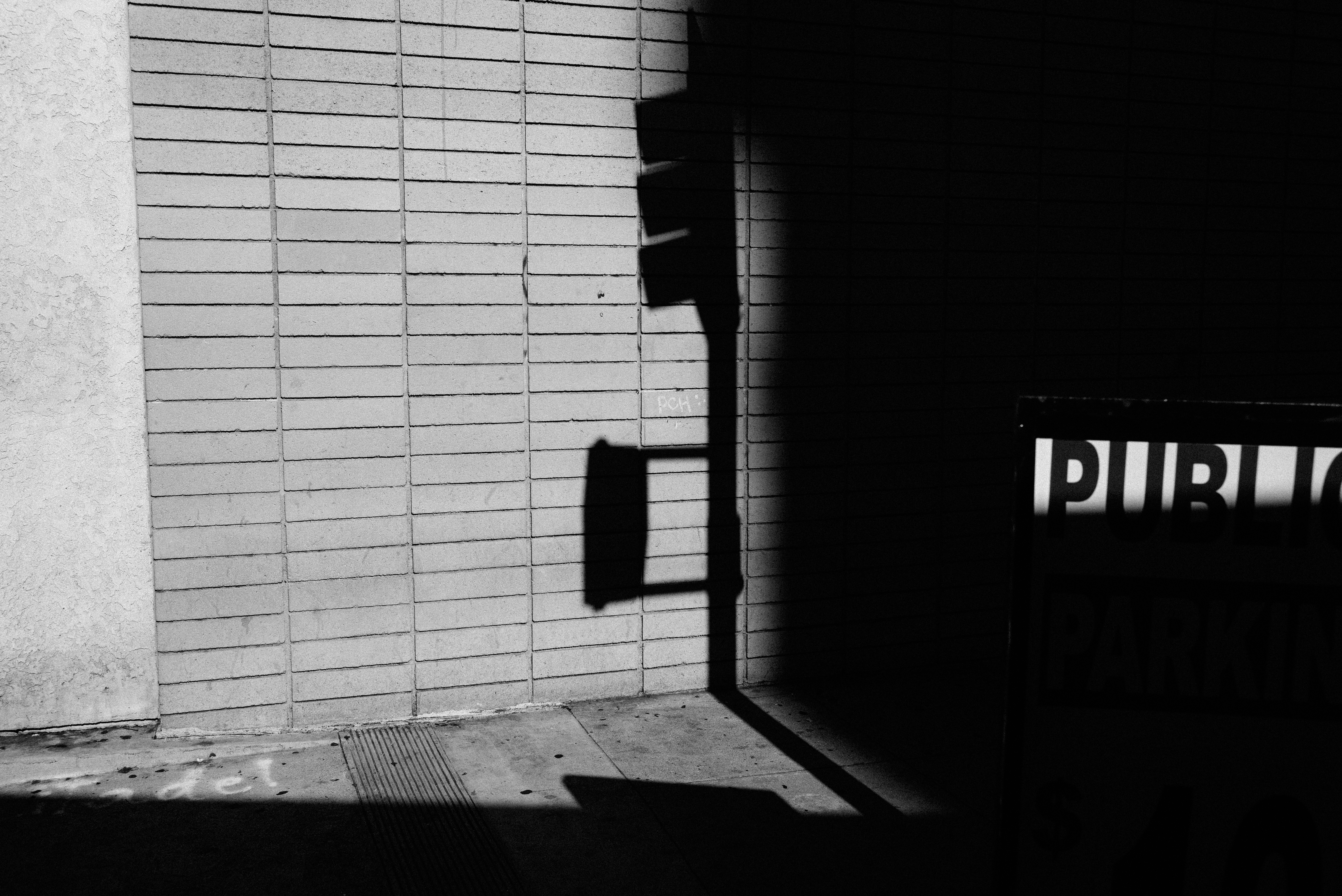 Diagonal sunlight and shadow cast across a brick wall and sidewalk with a silhouetted traffic walk sign.