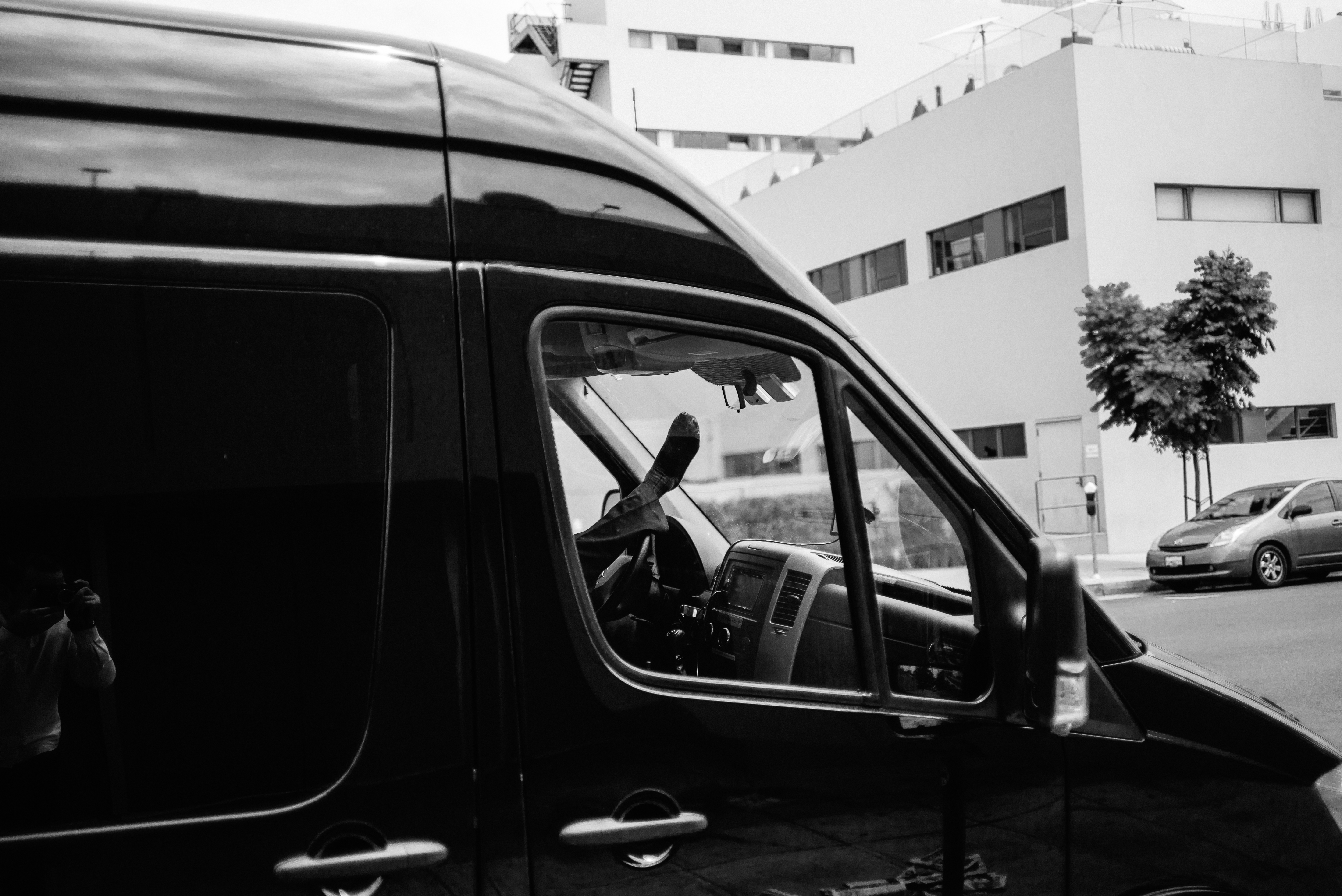 Person resting with a foot propped up on the dashboard of a black van along an urban street.
