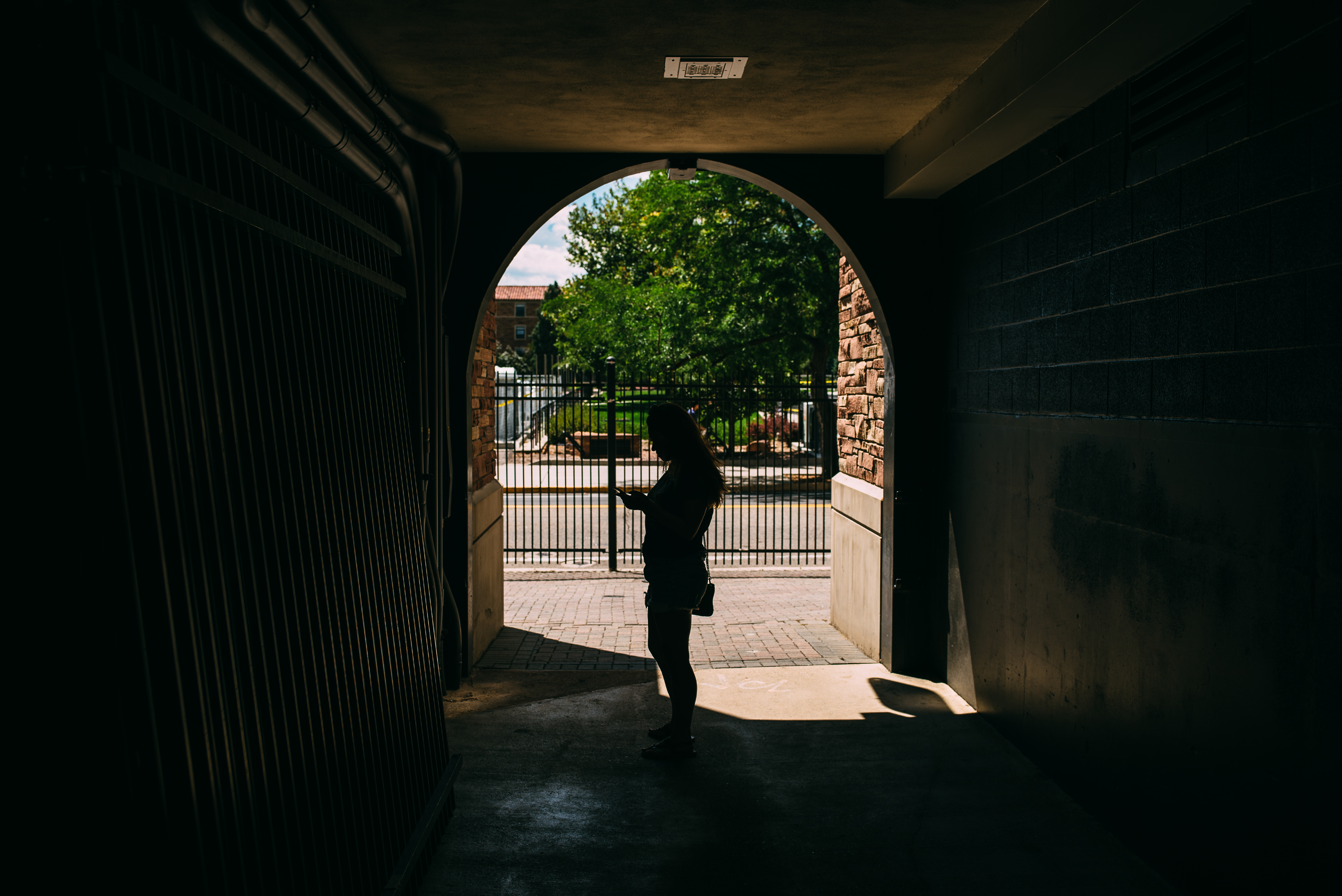 Person silhouetted inside a brick arch corridor checking a phone, with a sunlit courtyard outside.